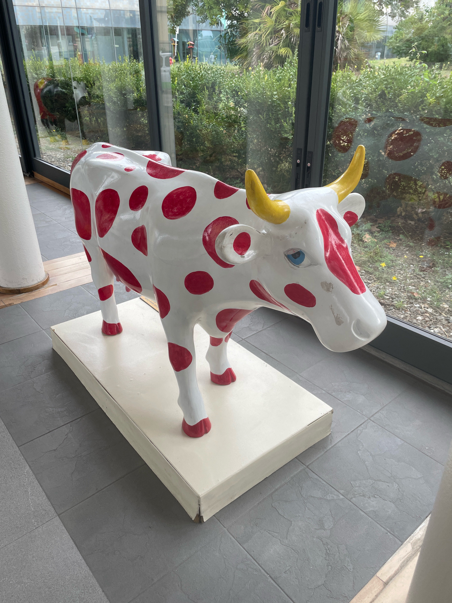 A life-sized, whimsical statue of a cow, painted white with bright red polka dots and yellow horns, stands on a cream-colored platform. The cow has blue eyes and appears to be in profile, facing right. It is situated indoors, next to a large window that looks out onto a lush green garden with trees and shrubs. The flooring in the foreground consists of large, grey tiles. The background, visible through the window, shows greenery and some architectural elements of a modern building. The overall scene suggests an art installation or decorative piece within a contemporary space, possibly a hotel, museum, or office building. The location is not Florence, Italy, but rather a nondescript indoor setting.