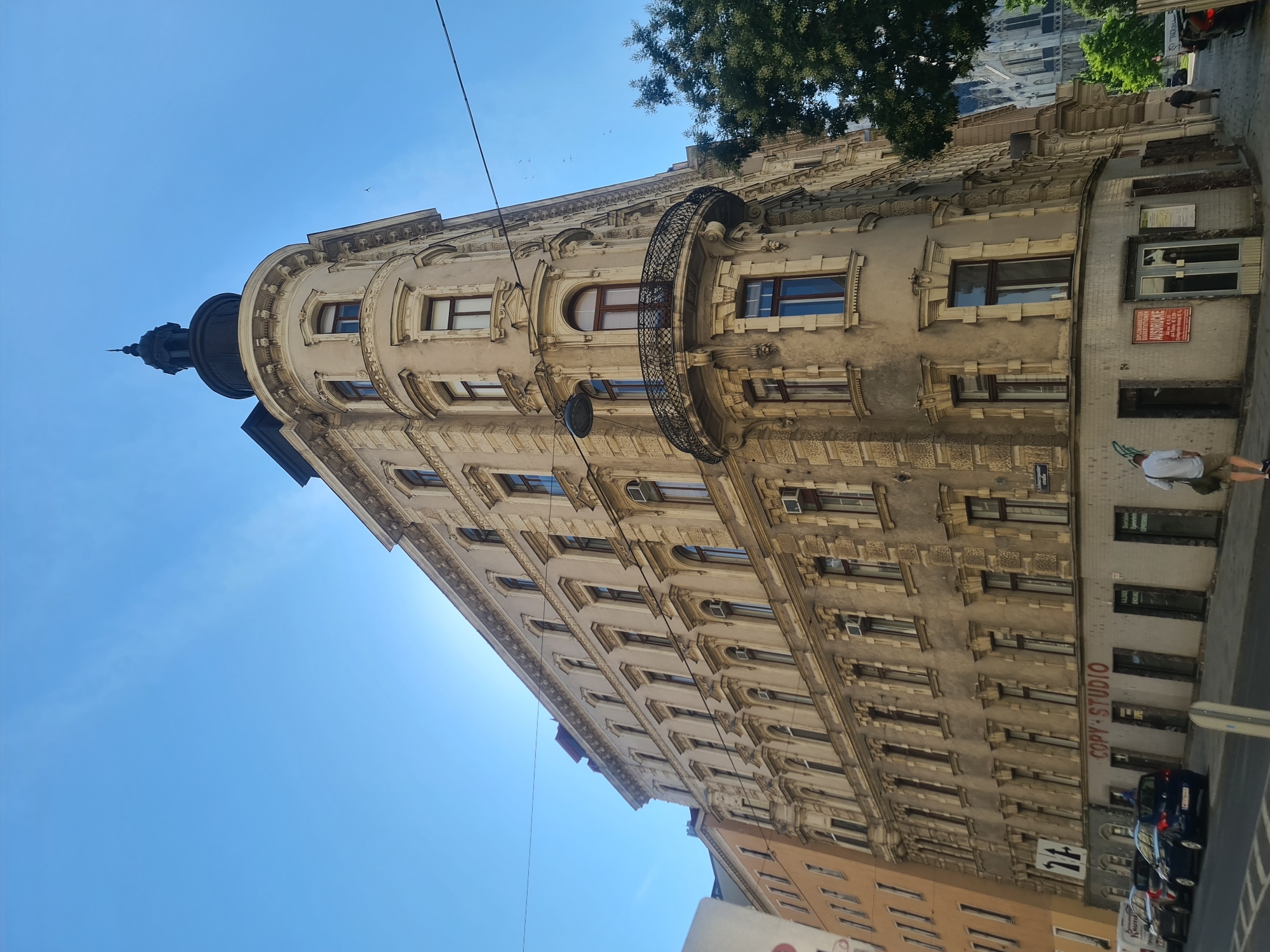 This is a low-angle, full shot of a multi-story building on a street in Vienna, Austria, under a clear blue sky. The building is constructed in an ornate architectural style, with light beige stucco facades, dark-framed windows, and decorative stone carvings around the upper floors. The building has a curved corner that extends upwards to a dark, domed turret with a spire. A wrought-iron balcony with an ornate railing is attached to the curved corner on one of the upper floors.
Below the building, on the street level, are storefronts. One storefront has red lettering that reads "COPY STUDIO". Other storefronts have various signs, including one that says "DIPLOMARBEITEN MASTERARBEITEN DISSERTATIONEN AUSDRUCKE" with pricing information. Several cars are parked along the curb on the street. A few people are visible on the sidewalk. One person, wearing a white shirt and khaki shorts, walks away from the camera towards the right. Another person, dressed in dark clothing, walks away from the camera towards the left. Overhead, power lines are strung across the sky. To the right of the building, a large green tree is visible, and in the background, a gothic-style church steeple can be seen. The overall impression is a daytime street scene in a European city.