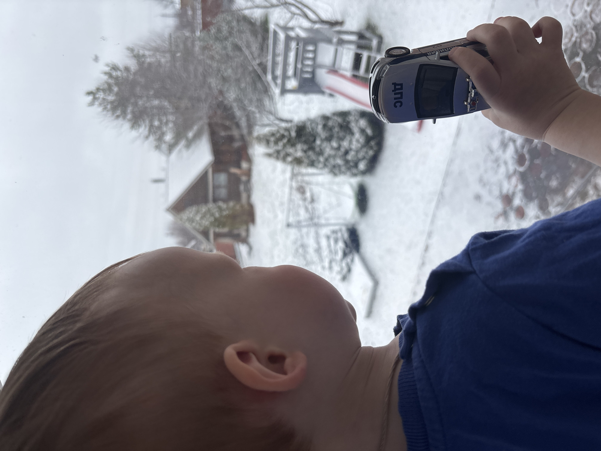 A child, wearing a blue shirt, is holding a toy car against a window. The reflection in the window shows a snow-covered yard with a house and a play structure. The toy car is silver and black and has the letters "ДПС" on it. The location is Ферма, Russia.