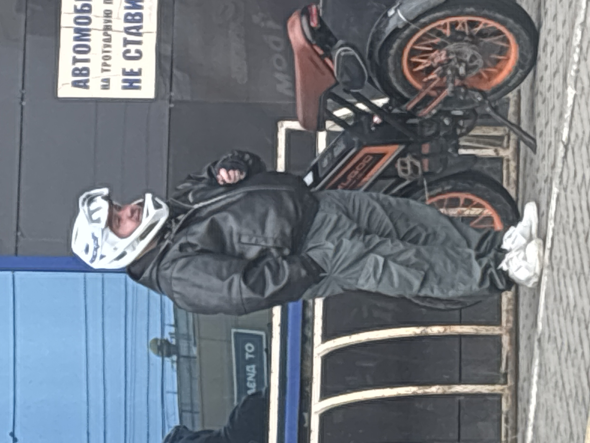 A man in a helmet and gray outerwear stands near a black and orange electric scooter on a city street. A sign with Russian text ("Автомобили на тротуарную не ставить") is visible in the background, indicating "Cars are not allowed on the sidewalk." Another sign with Russian text ("Лена то") is also visible. The location is likely Izhevsk, Russia. The pavement is grey and tiled.