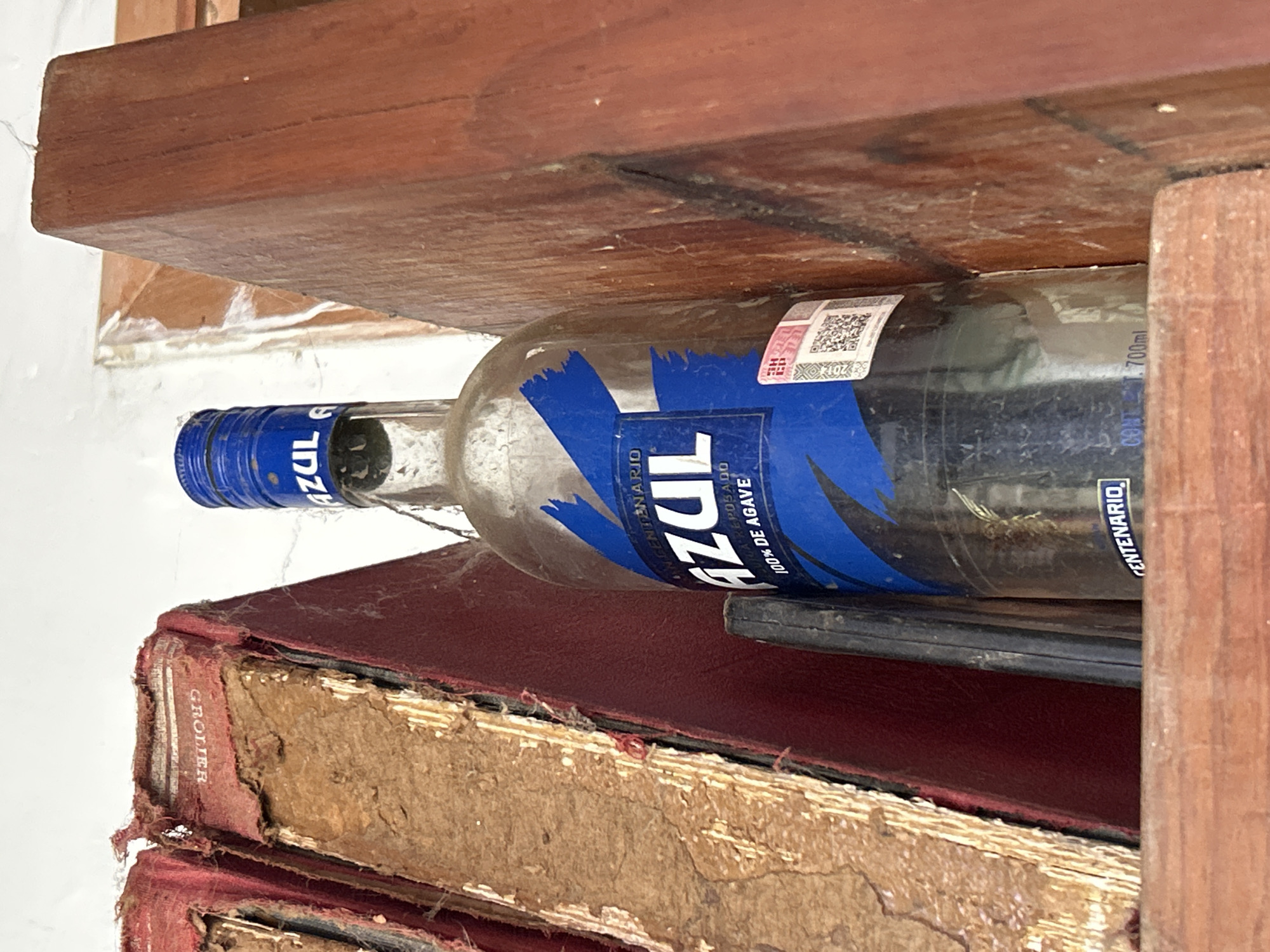 A close-up, low-angle shot shows a bottle of Centenario Azul tequila on a wooden shelf, wedged between old books and a wooden divider. The bottle is mostly empty, with a blue label featuring white lettering. A QR code sticker is visible on the label. The books, bound in red, appear aged and worn, with visible damage to their spines and pages. The wooden shelf and divider are made of dark wood, showing signs of wear. The background is a plain white wall, with some evidence of age and disrepair. The scene is dimly lit. The specific location in Santa Ana, Venezuela cannot be determined from the image.