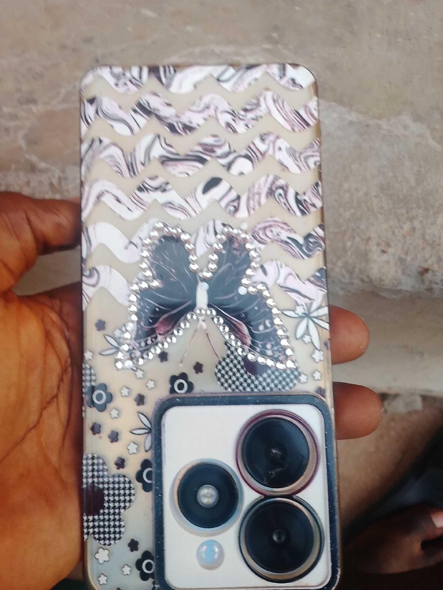 A hand holds a smartphone with a clear case featuring a white and black zigzag pattern at the top, a butterfly embellished with rhinestones in the center, and a floral pattern with black and white checkerboard flowers towards the bottom. The phone has a prominent camera module with three lenses. The background appears to be a rough, textured surface, possibly concrete or stone. The setting is outdoors, in an indeterminate urban location.