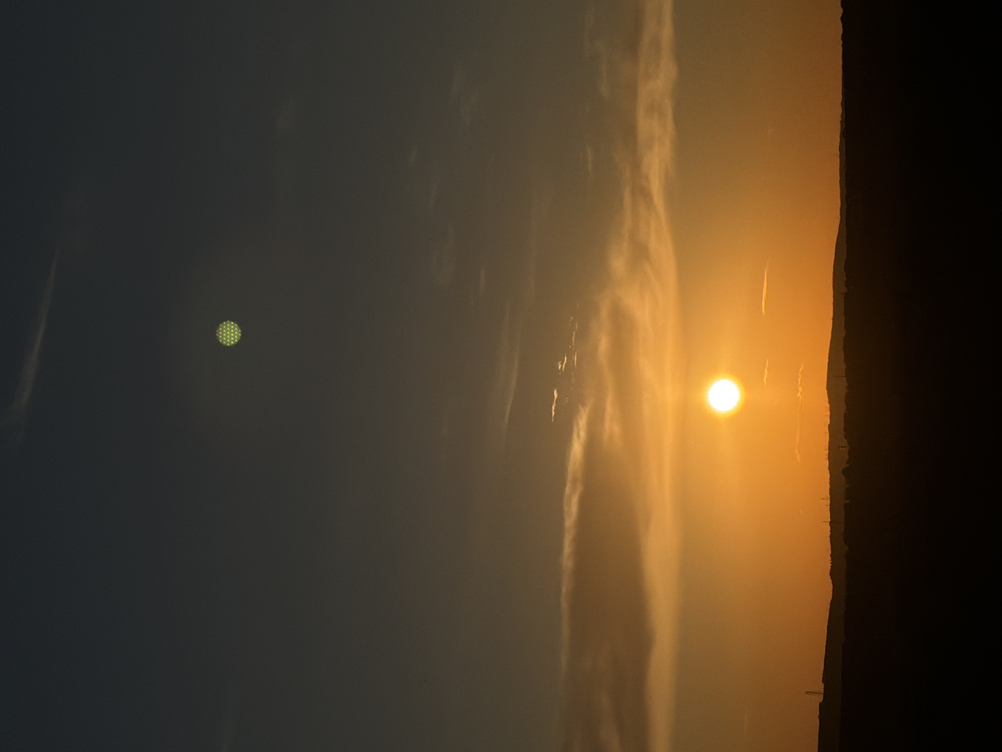 This is a vertical, medium shot photograph of a sunset over a rural landscape, likely near Stăuceni, Moldova. The sky transitions from a dark gray at the top to a bright orange at the horizon. Thin, wispy clouds are scattered throughout the sky, catching the warm hues of the sunset. In the lower portion of the image, a dark silhouette of rolling hills and sparse vegetation forms the horizon line. A bright, round sun is prominently visible near the center right of the frame, casting a strong orange glow. Above and to the left of the sun, a small, circular object with a distinct honeycomb pattern is visible in the darker sky. The lower horizon has a few faint vertical lines that might indicate distant structures or power lines.