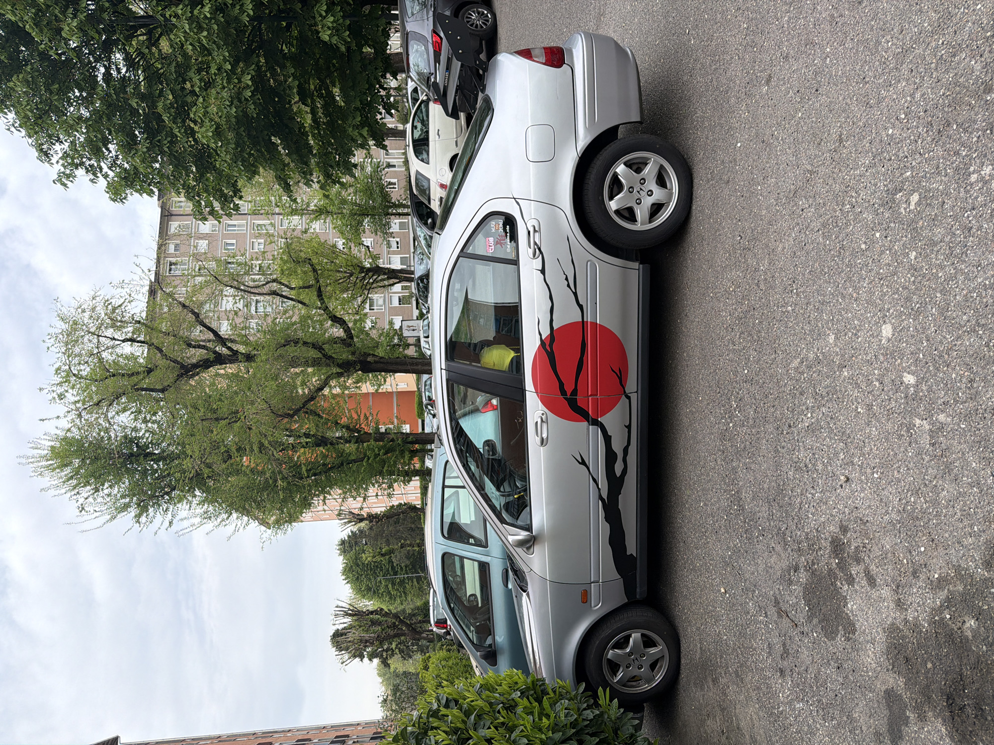 The image shows a silver four-door sedan parked on a paved street in Milan, Italy. The car features a large red circle with black tree branches decal on the side. The car is surrounded by other parked vehicles, trees, and bushes. In the background, there is a multi-story building. The environment suggests an urban or suburban residential area.