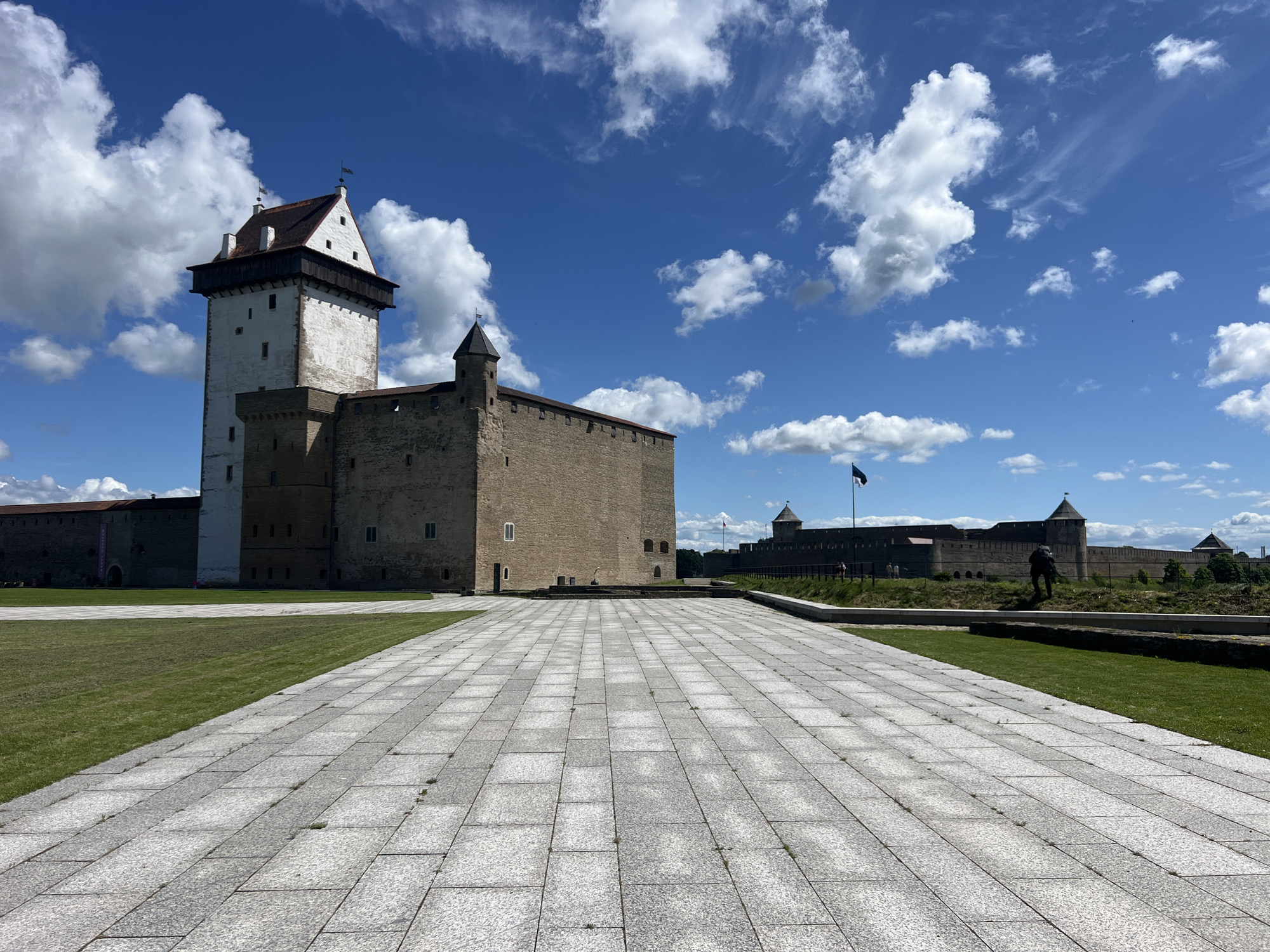 The image shows a wide view of Narva Castle in Narva, Estonia, on a sunny day with scattered clouds. In the foreground, a large paved area with gray stone tiles extends towards the castle. To the left of the paved area is a neatly mowed green lawn. The castle itself is a prominent feature, with a tall, white tower on the left, and a long, stone building with smaller turrets extending to the right. The castle walls are constructed of light brown stone. A dark flag flies from a pole on top of one of the turrets in the distance. The sky is a vibrant blue with fluffy white clouds. In the background, more of the castle's extensive fortifications can be seen. A lone figure is visible standing in front of the distant wall, appearing to look towards the castle. The overall scene depicts a historic landmark under a bright, clear sky, with a clean and well-maintained public space in front of it.
