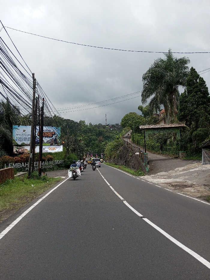 The image displays a two-lane asphalt road under an overcast sky in the city of Chendenbji, Bhutan. The road features solid white lines marking the shoulders and a broken white line as the center divider. On the left, multiple utility poles are densely wired with black cables, standing in front of lush green vegetation. Two large billboards are present; the leftmost advertises "LEMBAH CIBEUREUMAI RESORT" with contact details, while the rightmost displays a motorcycle and pricing information "40RB+" and "90K". A line of motorcycles, each carrying a rider, proceeds away from the viewer in the left lane. The foremost motorcycle is heavily laden with white sacks on its rear. A light-colored car is visible further down the road, also traveling away. On the right side of the road, dense green foliage, including a prominent palm tree, borders a concrete retaining wall. A green arched sign, partially obscured, marks an entrance to an ascending path on the right. A small, simple structure with a corrugated roof is positioned on the far right edge of the foreground. Hilly terrain covered in vegetation extends into the background, where scattered buildings and a communication tower are visible against the dense grey cloud cover.