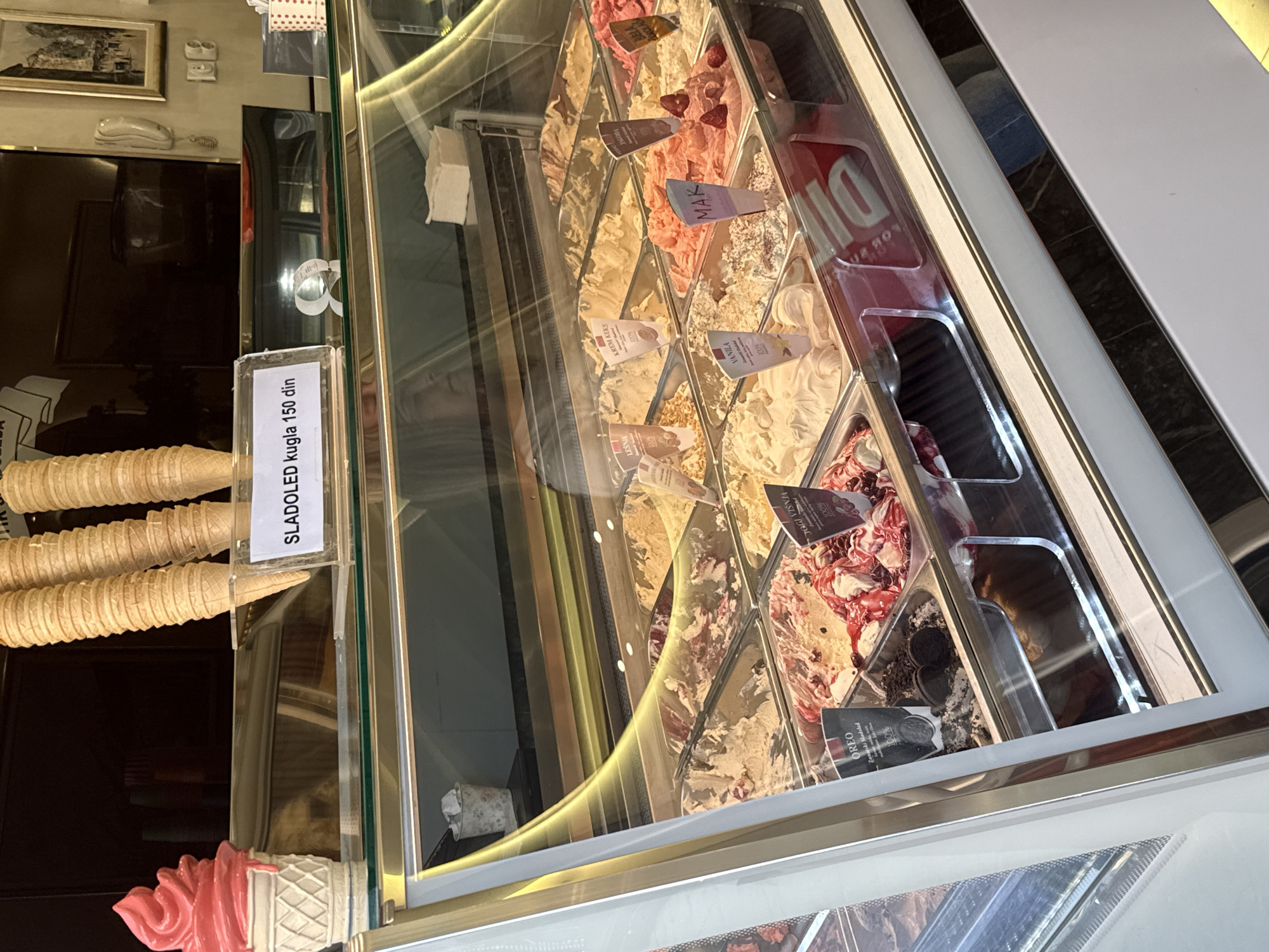 This image captures a close-up view of an ice cream display counter, likely in an ice cream parlor or cafe in Kragujevac, Serbia. The scene is indoors, brightly lit, showcasing a variety of ice cream flavors in metal trays beneath a clear glass cover.
Numerous flavors of ice cream are visible, ranging in color from creamy whites and browns to vibrant pinks and reds, some garnished with fruit pieces or cookie crumbles. Each flavor has a small plastic label identifying it. Visible labels include:
* "SLADOLED kugla 150 din" – indicating an ice cream scoop costs 150 Serbian Dinars.
* "JOGI VIŠNJA" – Yogurt Cherry (with a "100%" logo, implying natural ingredients).
* "OREO" – Oreo.
* "VANILA" – Vanilla (also with a "100%" logo).
* "KREM KEKS" – Cream Cookie.
* "LESNIK" – Hazelnut (also with a "100%" logo).
* "MAK" – Poppy Seed (partially visible).
* "JAGOD" – Strawberry (partially visible).
To the left of the display case, a stack of multiple waffle ice cream cones is neatly arranged, suggesting they are ready for use. Below the cones, a decorative, pink soft-serve ice cream cone model is visible. Reflections in the glass display case provide glimpses of the surrounding environment, including a framed picture on a wall, an old-fashioned white telephone, and faint, distorted reflections of what appears to be a person, possibly a customer or staff member, and other interior elements. The ambiance suggests a clean and inviting establishment.
