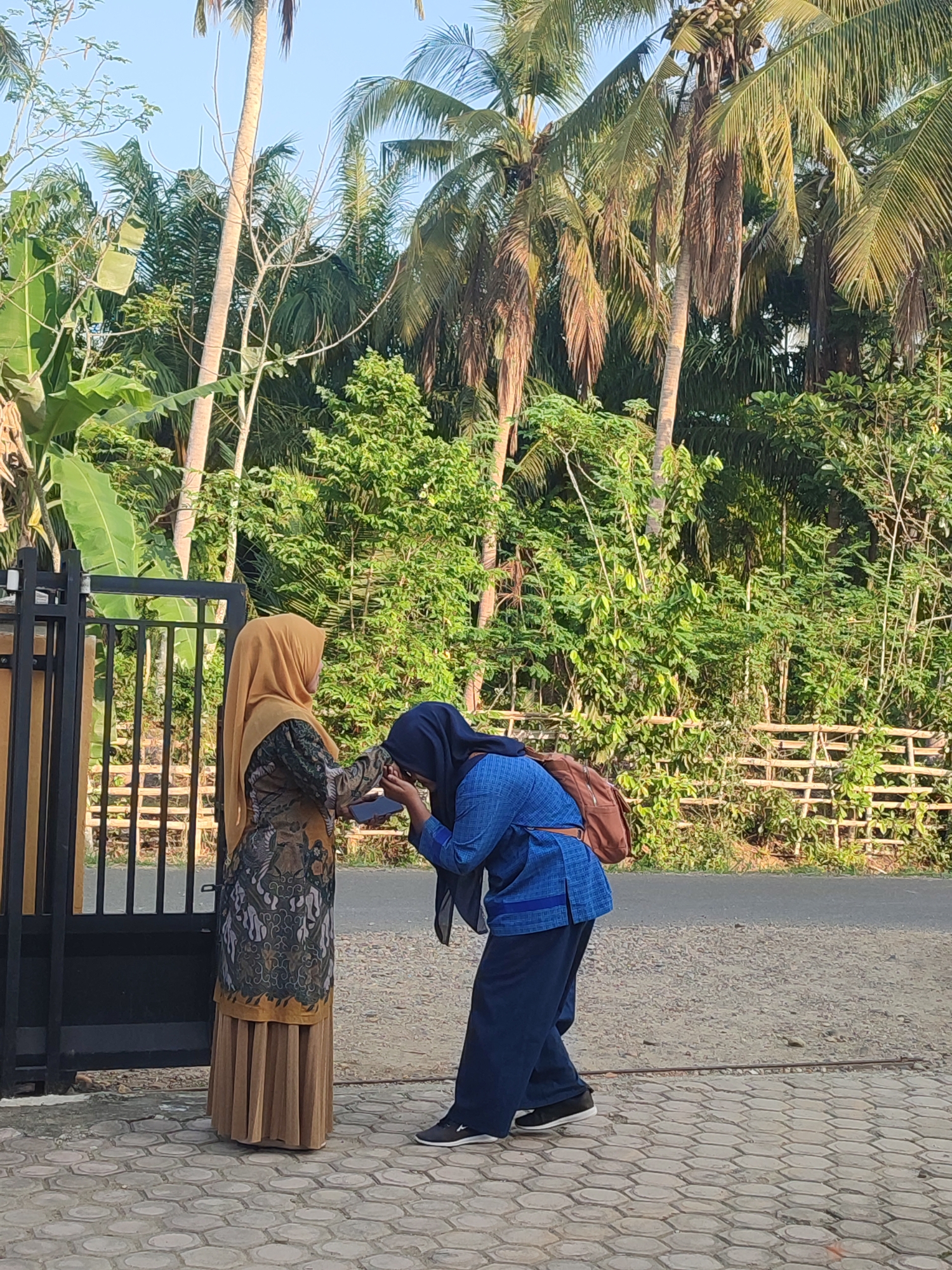 Two women stand near a gate on a paved surface in Blangriek, Indonesia. The woman on the left wears a tan headscarf, a patterned top, and a long tan skirt. The woman on the right wears a blue headscarf, a blue top, dark blue pants, and black sneakers. She also has a brown backpack. The woman on the right is bowing and touching the hand of the woman on the left. Behind them is a fence, lush green vegetation including palm trees, and a narrow road. The sky is visible above the trees.