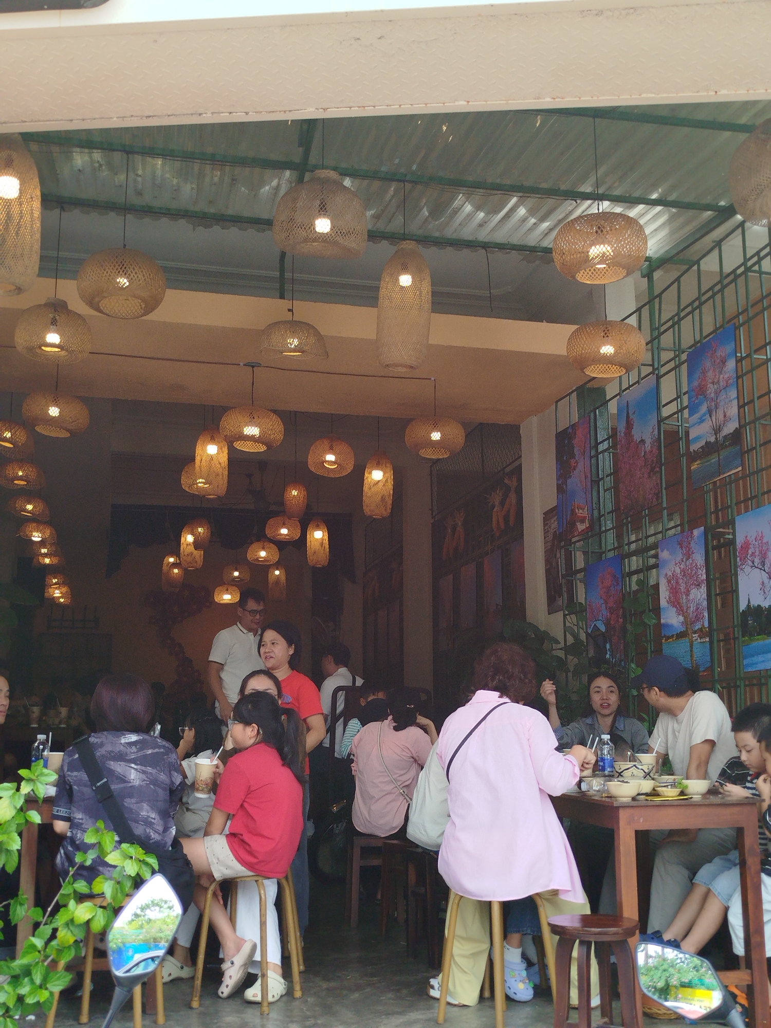 The image depicts an outdoor seating area of a restaurant in Da Lat, Vietnam, characterized by its abundant rattan pendant lights hanging from a corrugated metal roof. The atmosphere is lively, with several patrons seated at wooden tables, engaged in dining and conversation. In the foreground, a person in a dark patterned shirt is seated, facing away from the camera. To their right, a young girl in a red t-shirt and shorts is seated, looking to her left. Further right, a woman in a pink shirt and yellow pants is seated at a table, interacting with a man wearing a blue cap and a white t-shirt. Another man in a white t-shirt stands behind the woman in the pink shirt. A young boy in a striped shirt is visible on the far right, seated at a table with adults. The walls of the establishment are decorated with colorful artwork featuring trees and landscapes. Green foliage is visible in the lower left corner, and a motorcycle mirror reflects a green landscape. The overall scene conveys a casual dining experience within a visually appealing, naturally lit environment.