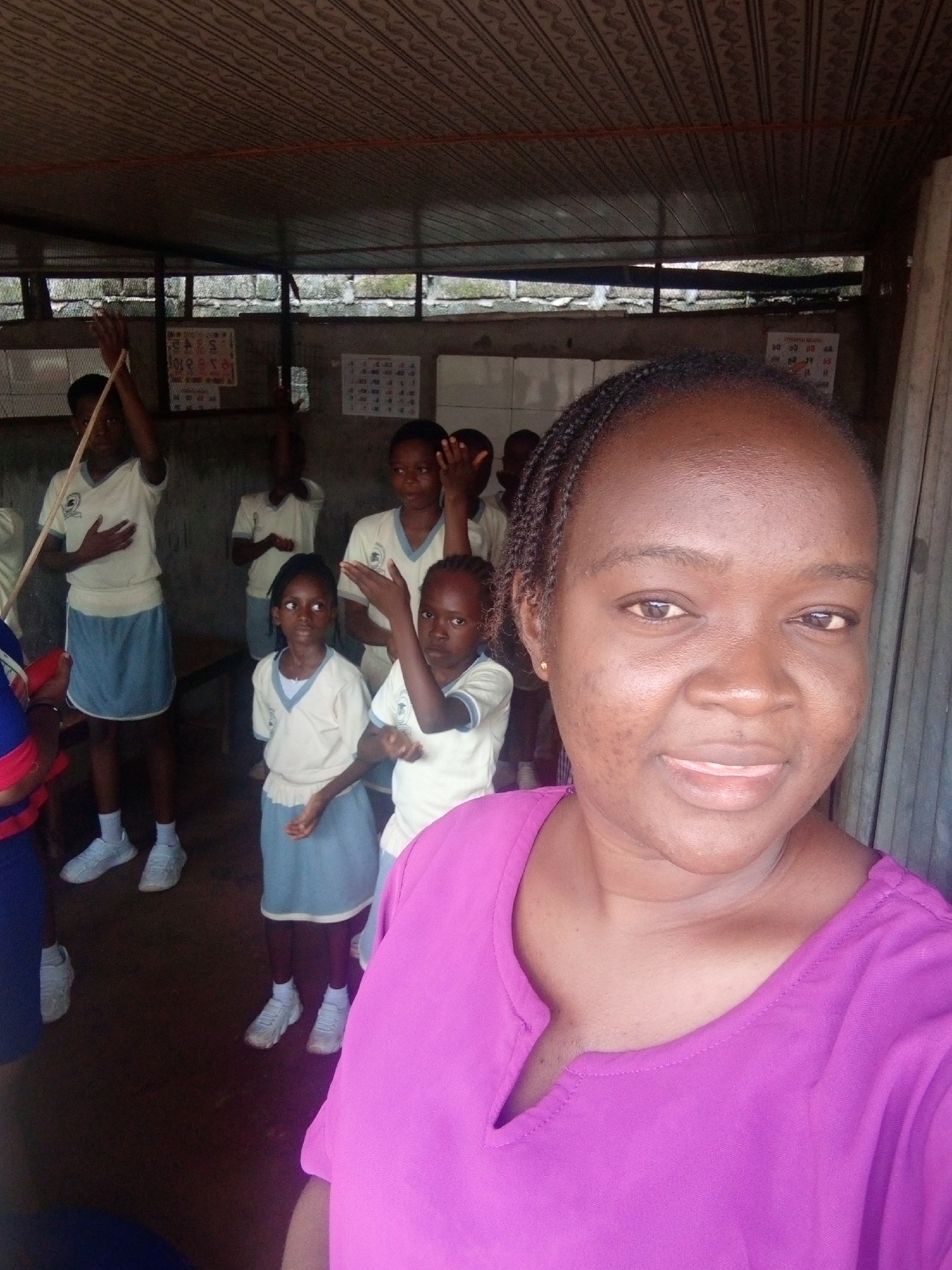In Ukwu-Abwa, Nigeria, a group of young students in school uniforms, consisting of white tops with blue trim and light blue skirts or shorts, stand indoors, possibly in a classroom or communal area with a corrugated metal roof and rough concrete walls. Some students are raising their hands, suggesting an activity or lesson is taking place. The foreground is dominated by a close-up of a woman wearing a vibrant purple top, her face turned towards the camera in a friendly smile. The background is partially obscured by the woman, but the children and the rustic environment of the building are visible.