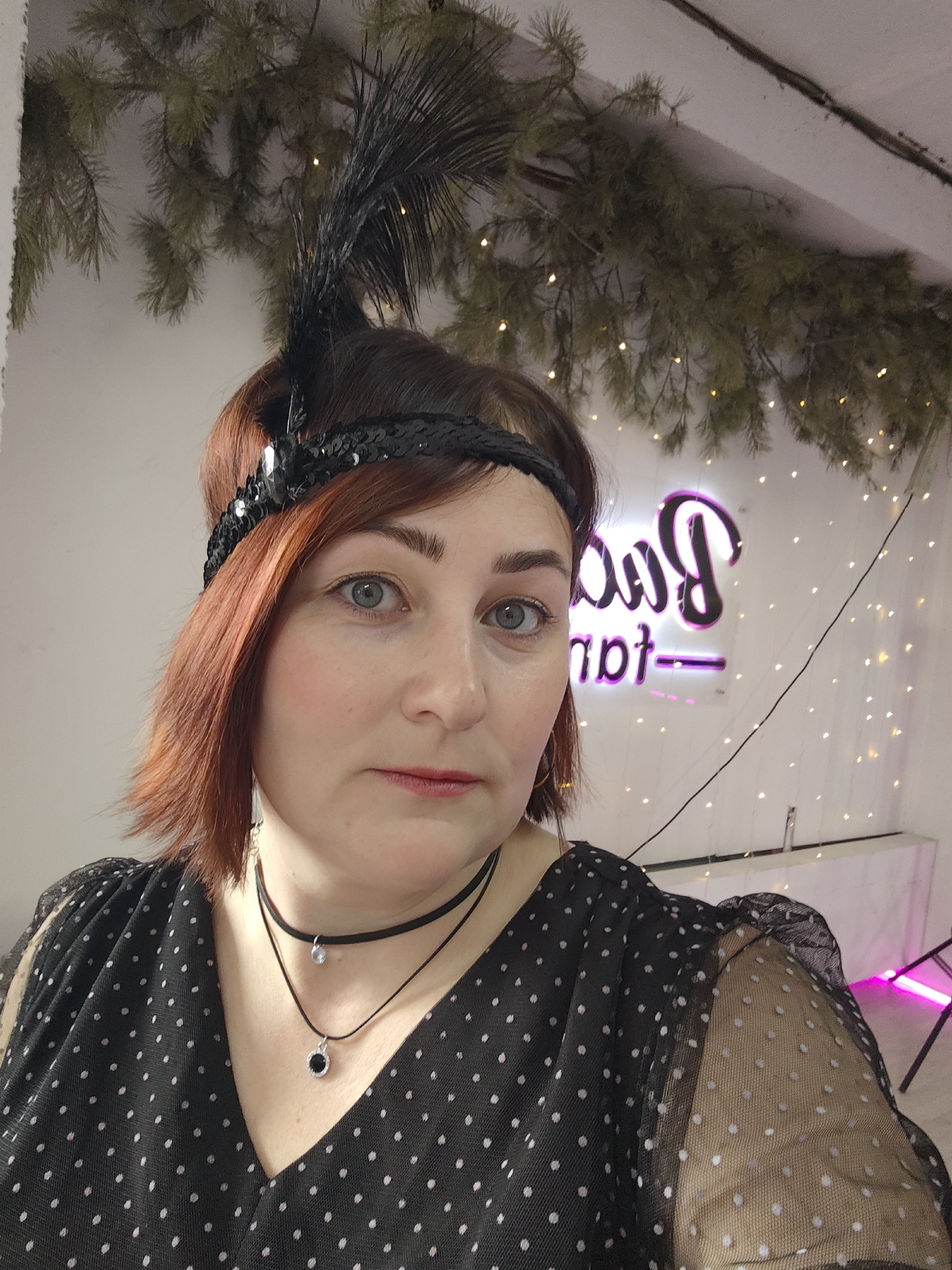 The image depicts a female individual taking a selfie indoors in Chaikovsky, Russia. The adult female subject has reddish-brown, medium-length hair and light-colored eyes, and is looking directly at the camera. She wears a black sequined headband with a black feather plume positioned on the viewer's left side of her head. Her attire includes a black garment with white polka dots, featuring a V-neckline and sheer, polka-dotted sleeves. She wears two necklaces: a black choker with a small, light-colored pendant, and a longer black cord necklace with a circular black pendant. The background consists of light-colored walls adorned with suspended evergreen branches and numerous warm-toned string lights. To the subject's right, a purple neon sign is partially visible, displaying stylized Cyrillic script, possibly "Баб" followed by "грt-". Below the neon sign, a white rectangular surface, potentially a table or counter, is present, along with a black stand emitting a pink glow further to the right. A thin black wire runs diagonally across the upper right portion of the background.