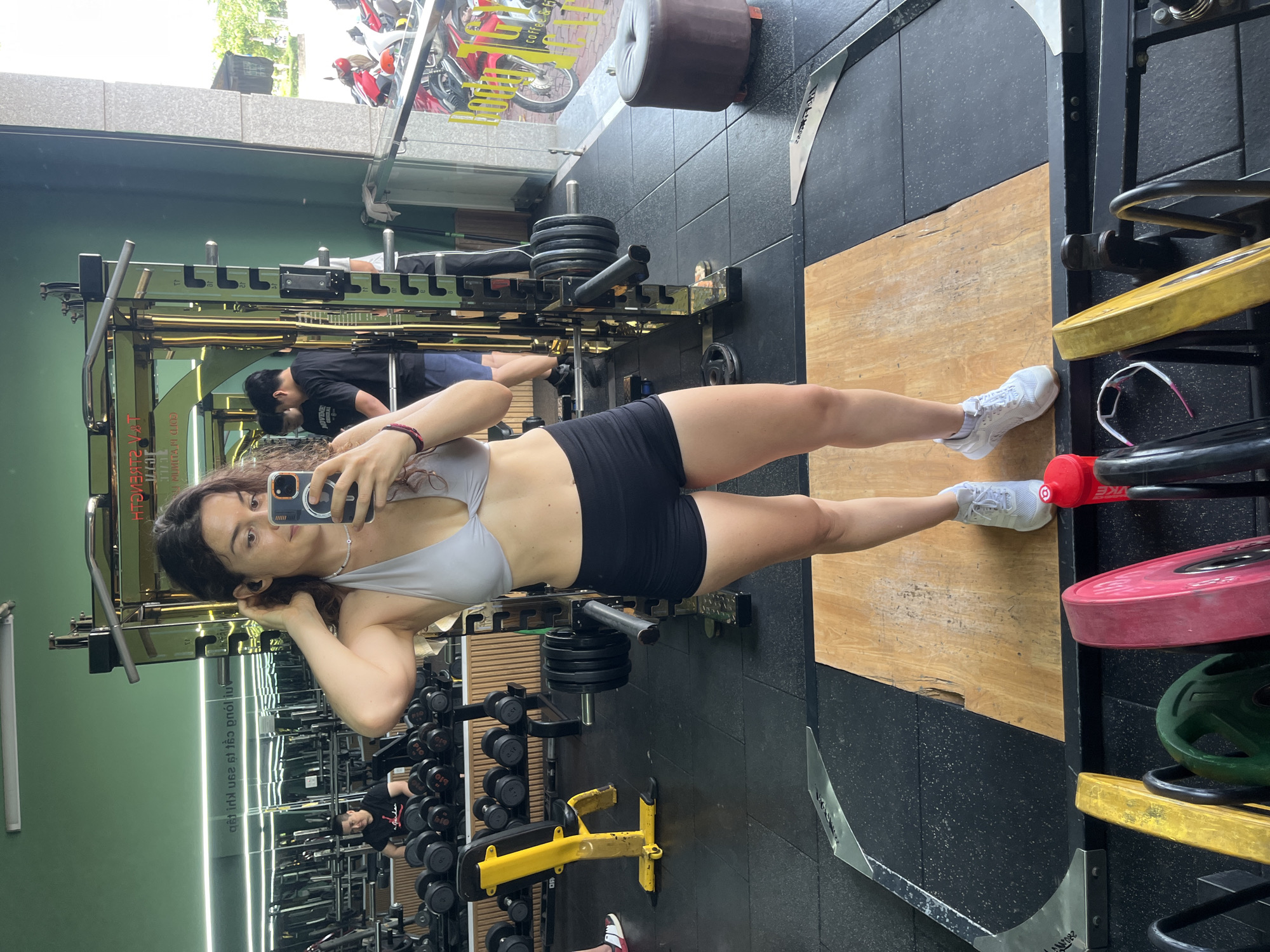A young woman with long, dark curly hair stands in a gym, taking a mirror selfie. She is wearing a light grey sports bra and black athletic shorts, along with white sneakers. Her right hand is touching her hair, and she is holding her phone in her left hand. In the background, a weight rack with barbells and weight plates is visible, as well as other gym equipment and people working out. Outside the gym, through a large window, motorcycles and a building are visible. The gym appears to be located in the city of Phú Thủy, Vietnam, as suggested by the text on the wall behind the woman, which reads "NƠI LUYỆN TẬP CHUYÊN NGHIỆP VIET GYM" and "THUỴ".