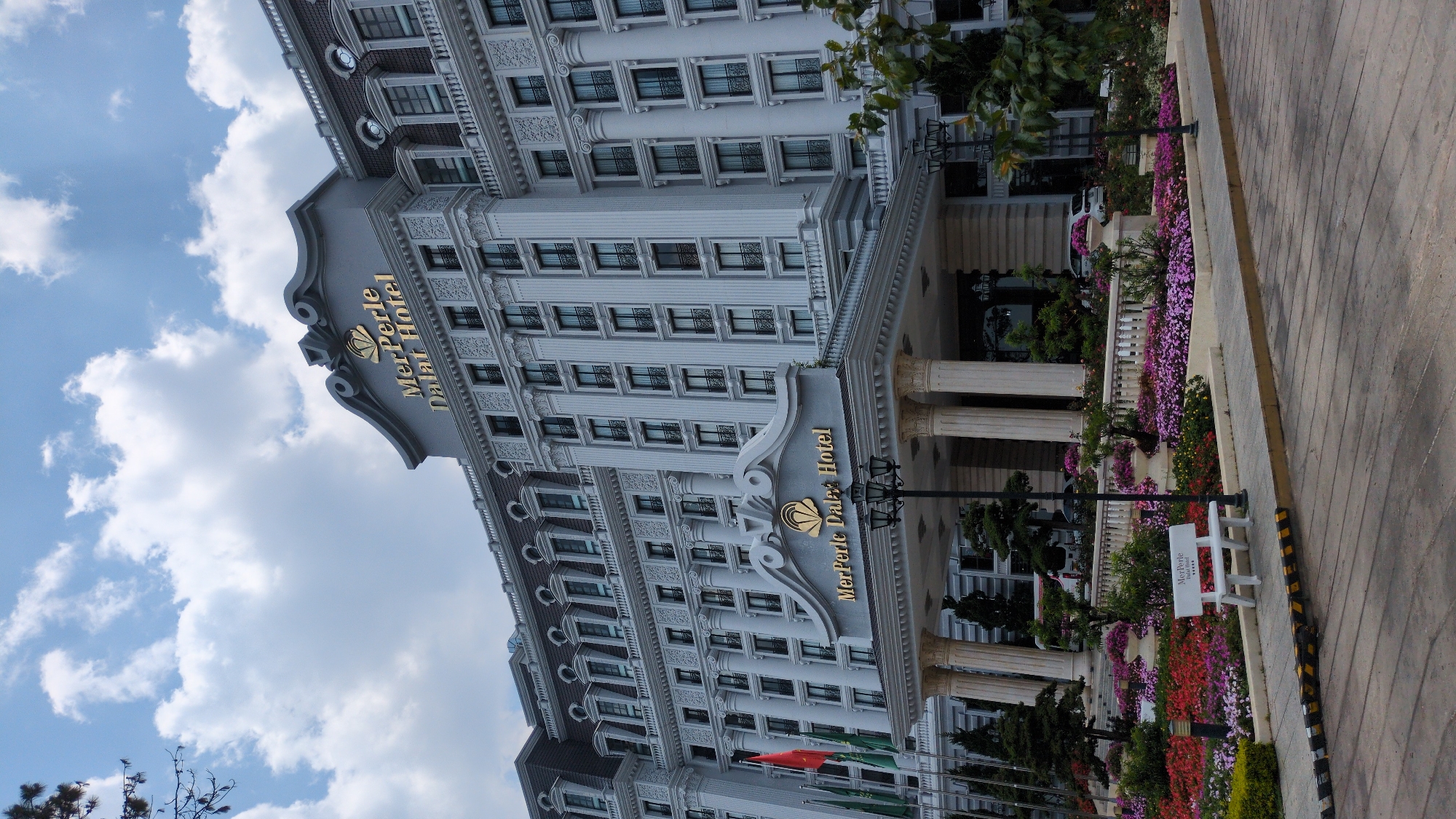 The image shows the MerPerle Da Lat Hotel in Da Lat, Vietnam. It's a grand building with multiple stories, ornate detailing, and many windows. The hotel's name is displayed prominently in gold lettering on a grey facade. The lower levels feature columns, flags, and landscaped flower beds. A sidewalk with yellow and black markings is visible in the foreground. The sky is partly cloudy. A bench with the name of the hotel is also visible.