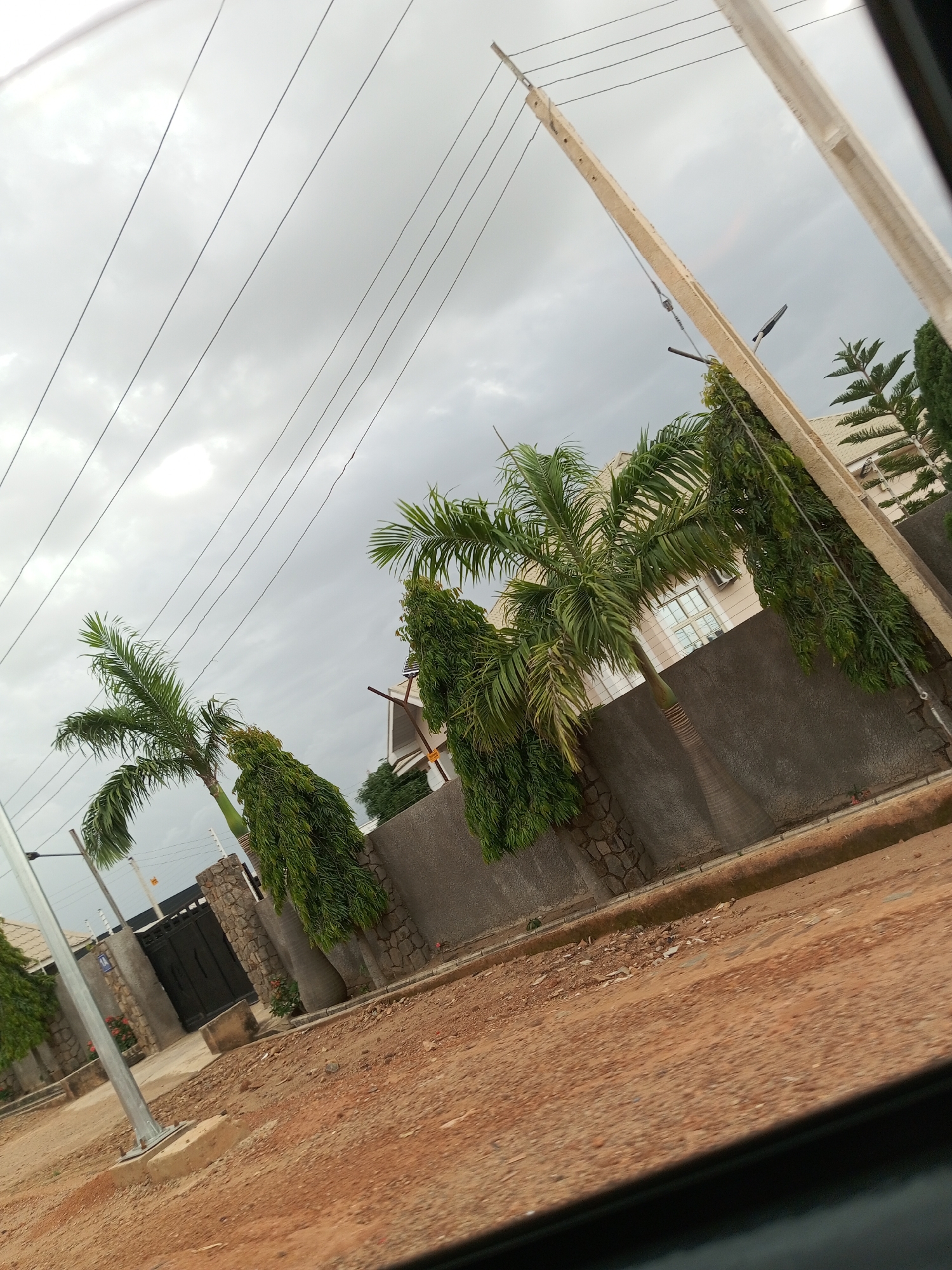 An outdoor, eye-level, full shot shows a view of a street with a building and trees. The scene is overcast, with gray clouds filling the sky. Power lines are strung diagonally across the upper portion of the frame, supported by telephone poles. In the middle ground, a stone wall lines the property, behind which are several trees, including palm trees and tall, slender trees. A multi-story building with cream-colored siding and windows is visible behind the trees. In the foreground, a dirt road with some scattered debris is present. To the left of the frame, a black metal gate is visible, set into the stone wall.