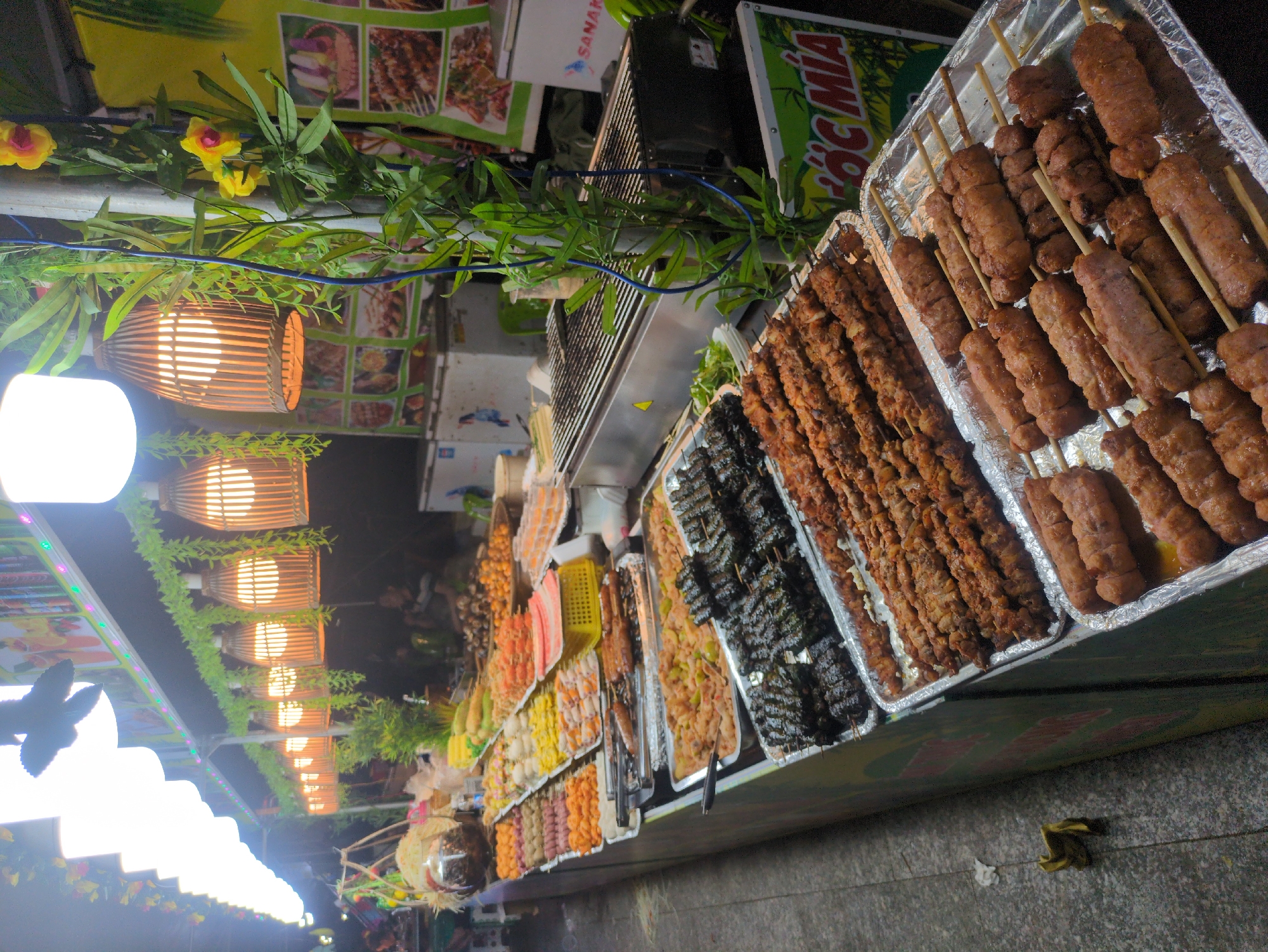 The image shows a street food stall in Tuy Hòa, Vietnam, featuring an array of grilled skewers on display. Various types of meat, seafood, and vegetable skewers are arranged in metal trays on a table. A grill can be seen behind the display, suggesting the food is prepared on site. The stall is illuminated by a row of hanging lights, some encased in bamboo shades, decorated with artificial flowers and greenery. The scene captures the lively atmosphere of a Vietnamese street food market.
