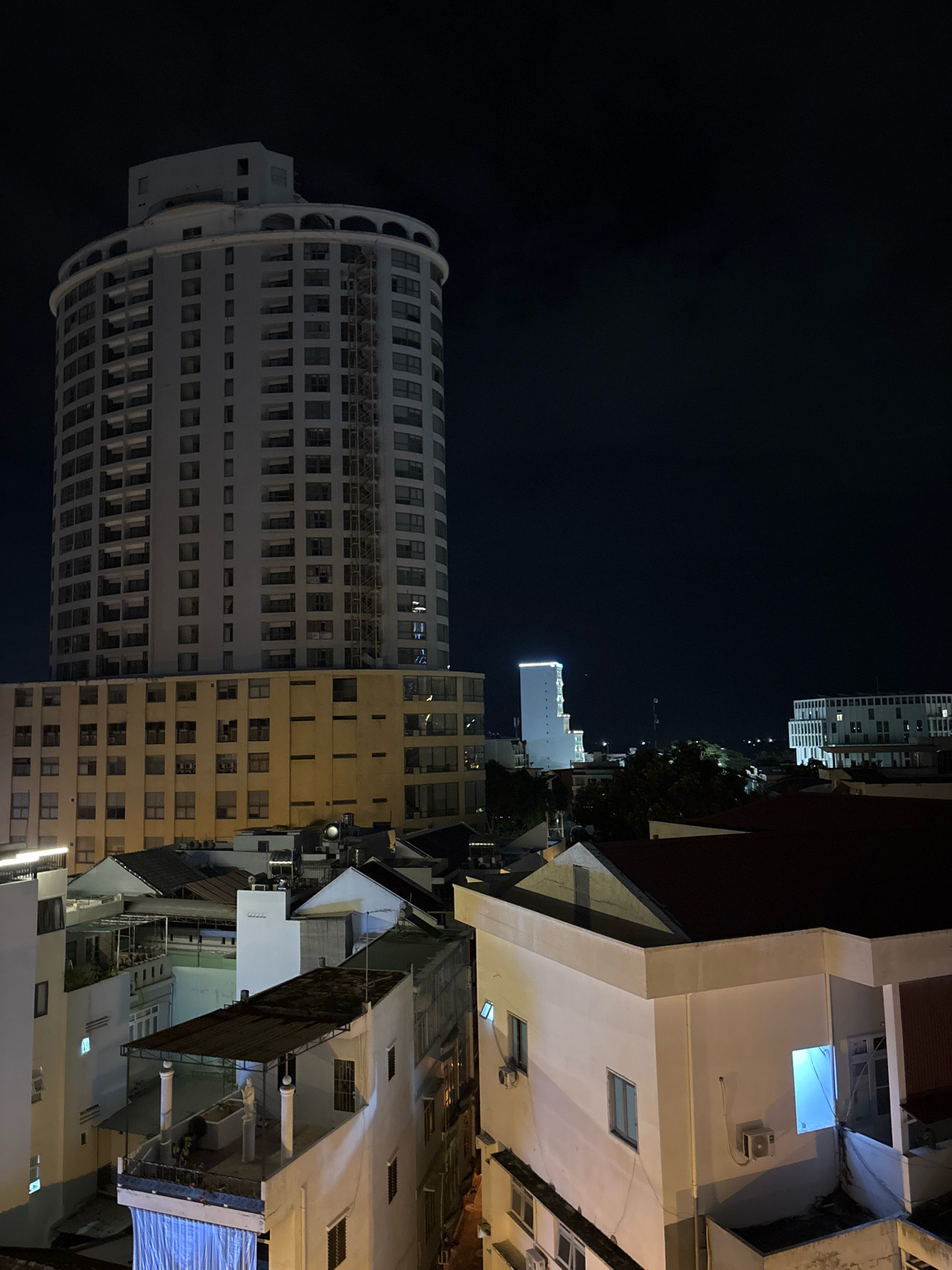 The image captures a nighttime view of an urban landscape in Nha Trang, Vietnam. A prominent, multi-story circular building with a white facade and arched balconies dominates the left side of the frame. Below this tower, a cream-colored building with numerous windows is visible, extending outwards. The foreground features several smaller buildings with various rooflines, some appearing residential with balconies and air conditioning units. A stark blue light emanates from a window on one of the closer buildings on the right. The sky is dark, indicating the late hour. The scene depicts a densely built urban environment with a mix of modern and older structures.