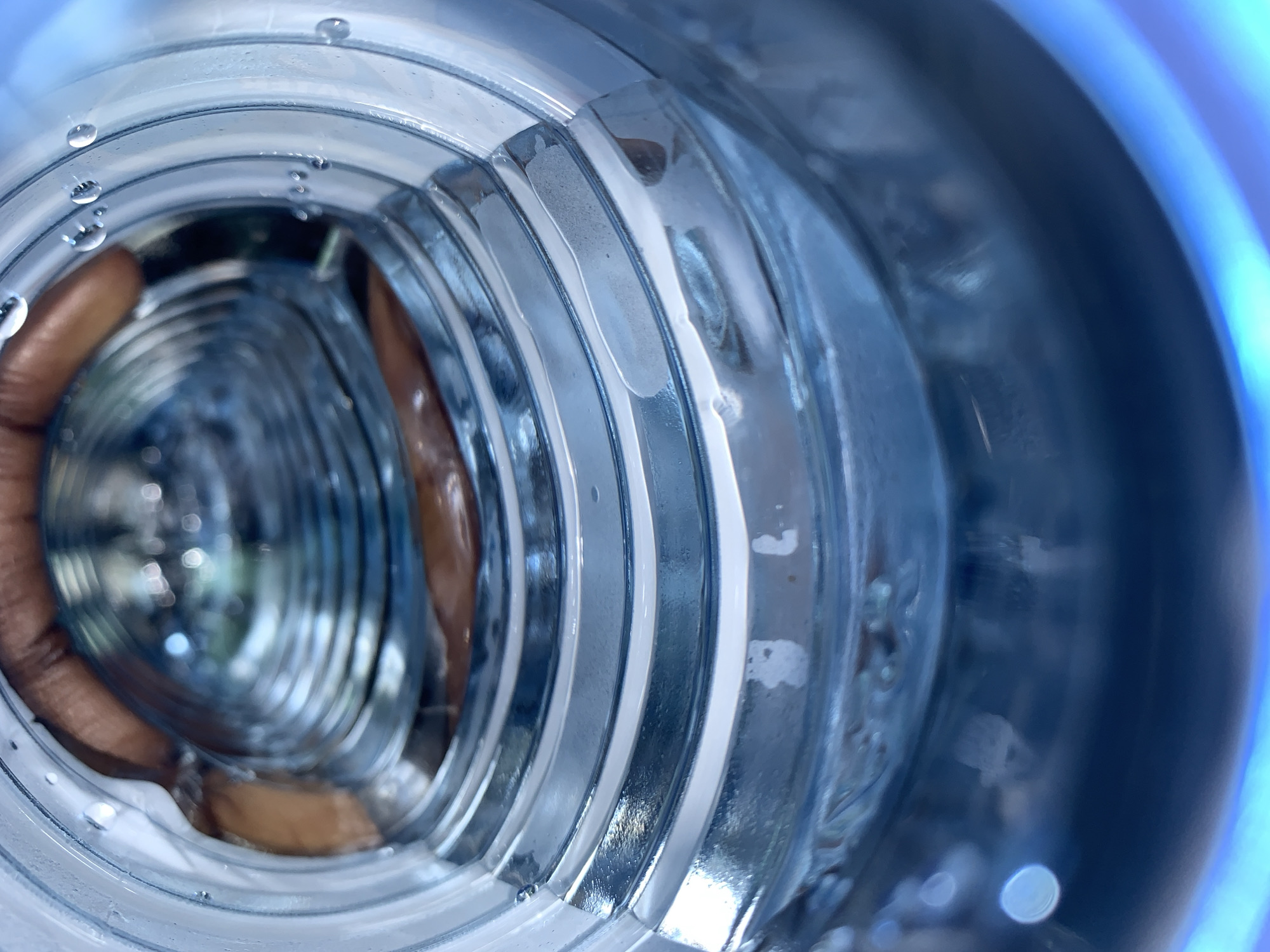 The image provides an internal, downward-looking perspective from the opening of a clear plastic bottle. The transparent internal walls of the bottle display a distinct pattern of concentric horizontal ribs or ridges. Small, spherical water droplets adhere to various points along the inner surface of these plastic walls. Further down the bottle's length, obscured by significant blurring and distortion, are reddish-brown forms that are consistent with human fingers or a hand wrapped around the exterior of the bottle. The lowest visible area within the bottle is characterized by a deep blue coloration, which may stem from a liquid contained within or a powerful blue light source reflecting off the bottle's base. Scattered, circular out-of-focus light reflections, known as bokeh, are also present in the deeper, blurred regions of the image. This scene is situated in the city of New Stadium, Nigeria.