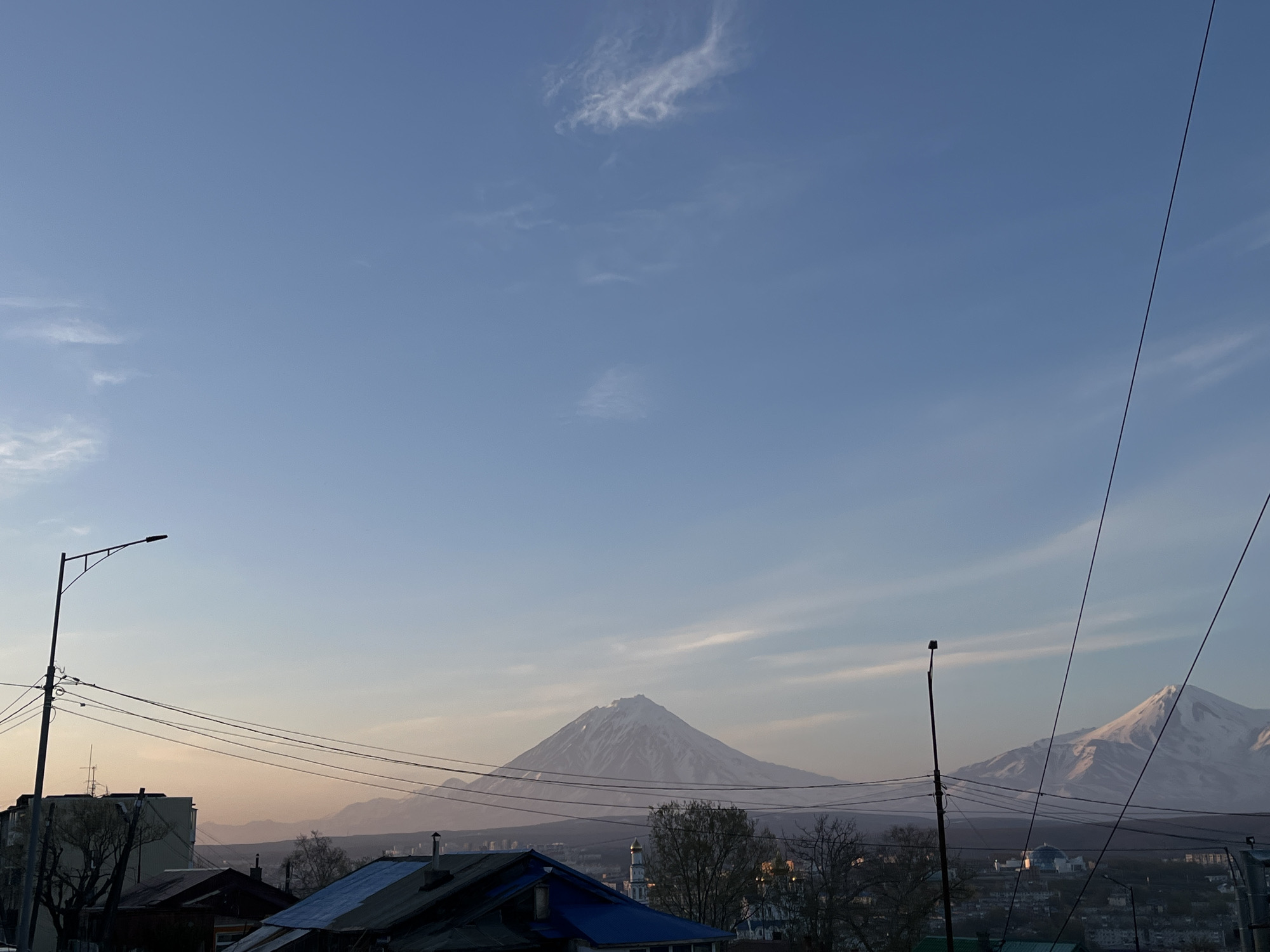 The image captures a panoramic view of a city landscape dominated by prominent snow-capped mountains under a clear blue sky. In the foreground, a network of utility poles and numerous power lines spans across the lower and mid-sections of the frame. A streetlight pole is visible on the far left.
Below the horizon, the rooftops of several buildings are seen, with prominent dark and blue-colored roofs. Further into the city, indistinct buildings and bare trees hint at an urban environment. Two specific structures stand out: one with a distinctive golden dome and another with a blue dome, suggesting architectural landmarks within the city.
The most striking features are the two majestic, snow-covered mountains in the distance. The central mountain is a classic conical peak, its slopes covered in white snow, while a broader, snow-dusted mountain rises to the right. The lower parts of the mountains and the horizon appear slightly hazy, indicating atmospheric depth.
The sky is a serene, vibrant blue, transitioning to a lighter, softer hue near the horizon. A few wispy white clouds are scattered sparsely across the upper sky. The soft, somewhat warm lighting on the mountains and the gentle gradient of the sky suggest either an early morning or late afternoon time of day. The presence of snow on the mountains indicates a cold climate. No people are visible, and there is no discernible text in the image. This landscape is characteristic of Petropavlovsk-Kamchatsky, Russia, known for its volcanic terrain.