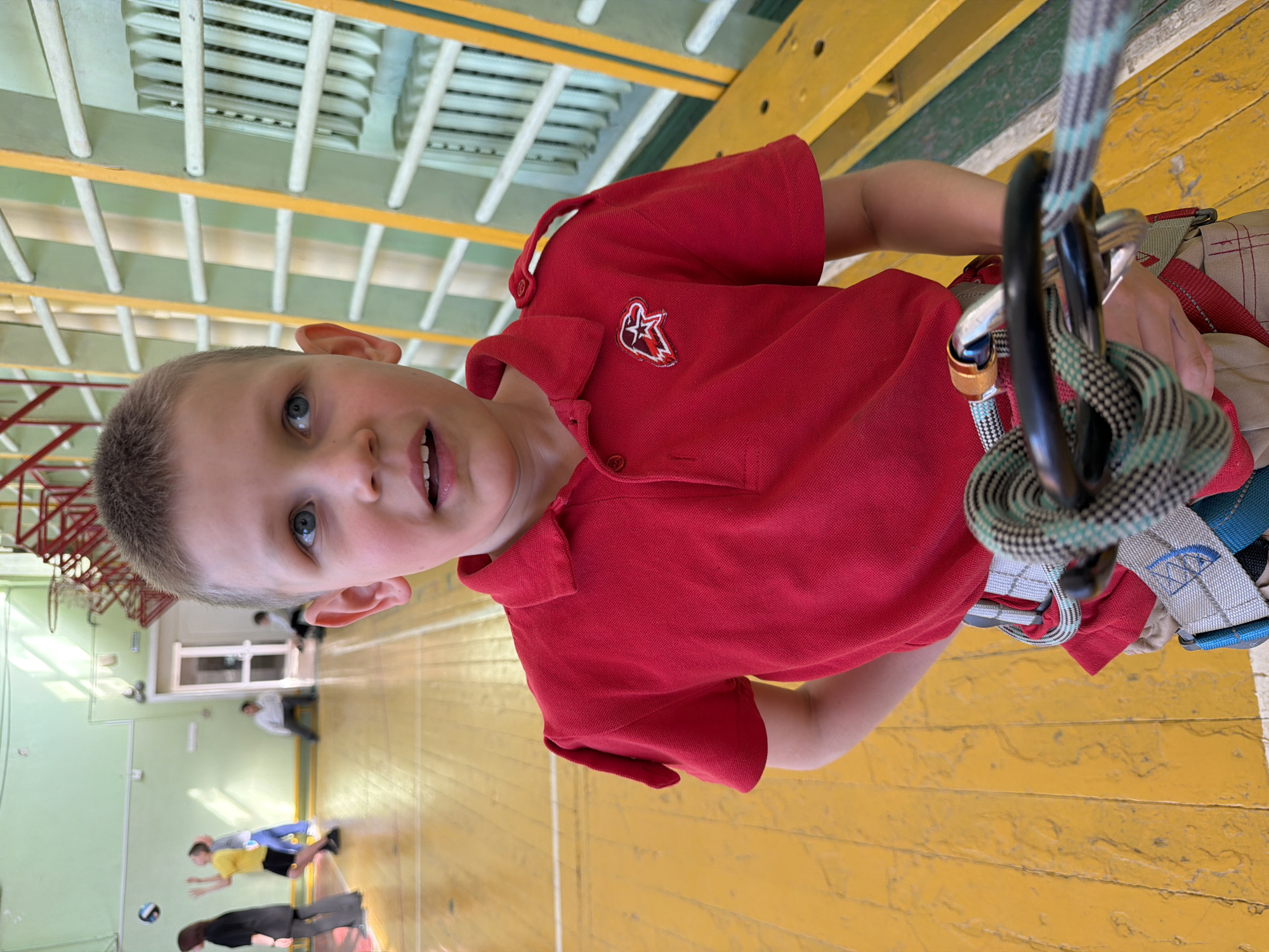 A young boy with short, light brown hair and blue eyes is harnessed and secured with climbing gear. He wears a red polo shirt with a logo on the left breast and looks towards the camera with a slight smile. The scene appears to be indoors, likely in a gymnasium, indicated by the wooden climbing wall he is attached to, as well as the sprung wooden floor and other gym equipment visible in the background. Other people are visible in the blurred background, some engaged in volleyball. The scene appears to be in Izhevsk, Russia.
