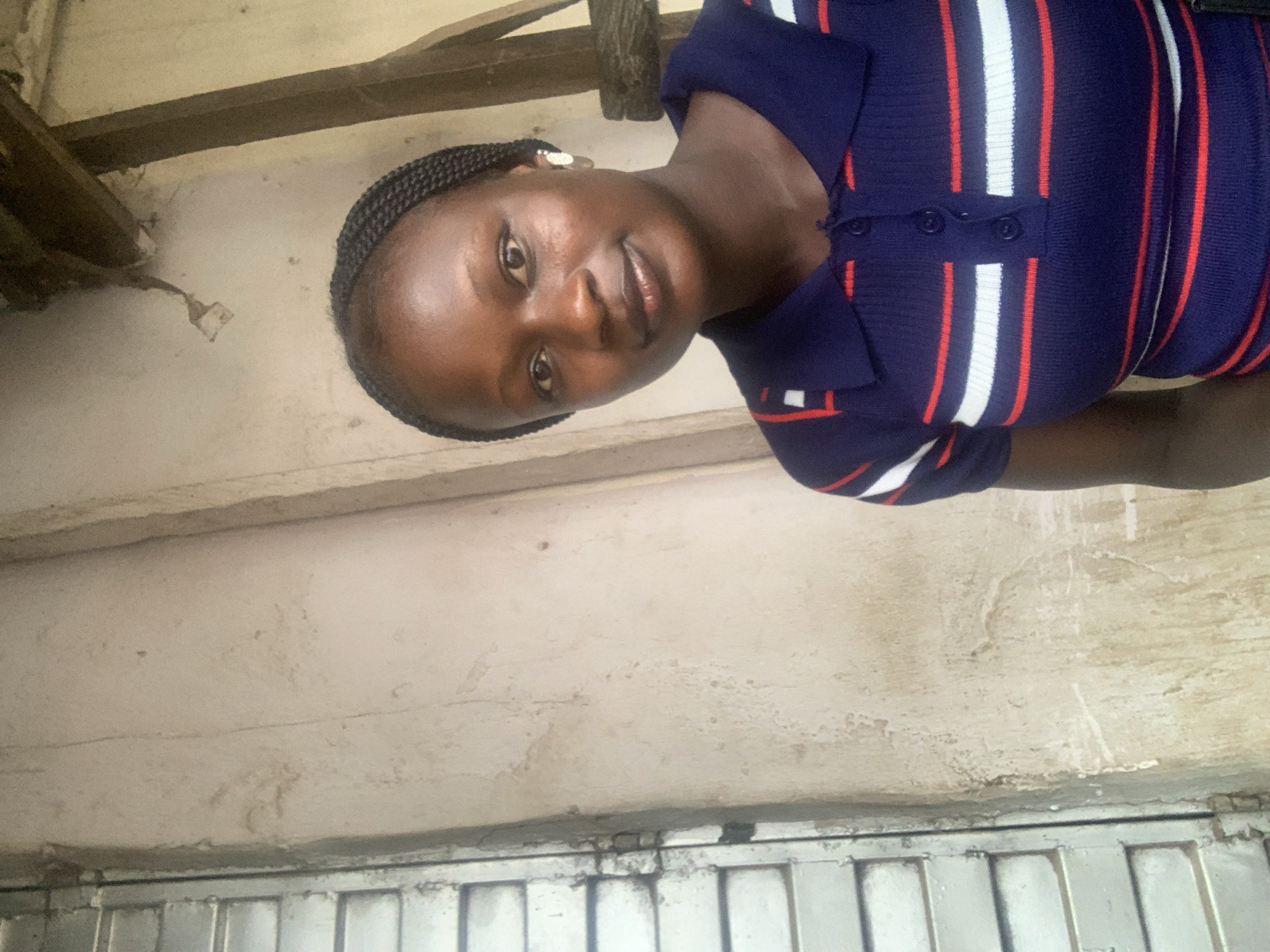 A young woman with braided hair stands indoors. She is wearing a navy blue, striped top with red and white stripes. The background includes a textured white wall, a wooden beam, and a metal garage door. The location of the photograph may be in Jos, Nigeria.