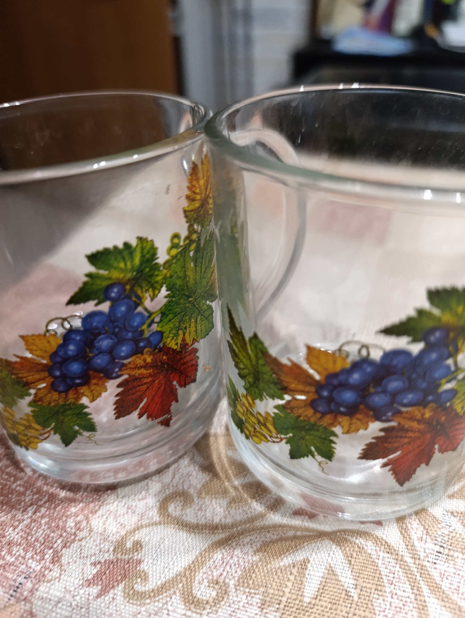 Two clear glass mugs decorated with a pattern of blue grapes and autumn leaves are placed side-by-side on a patterned tablecloth. The mugs are empty and the background is blurred, suggesting an indoor setting.