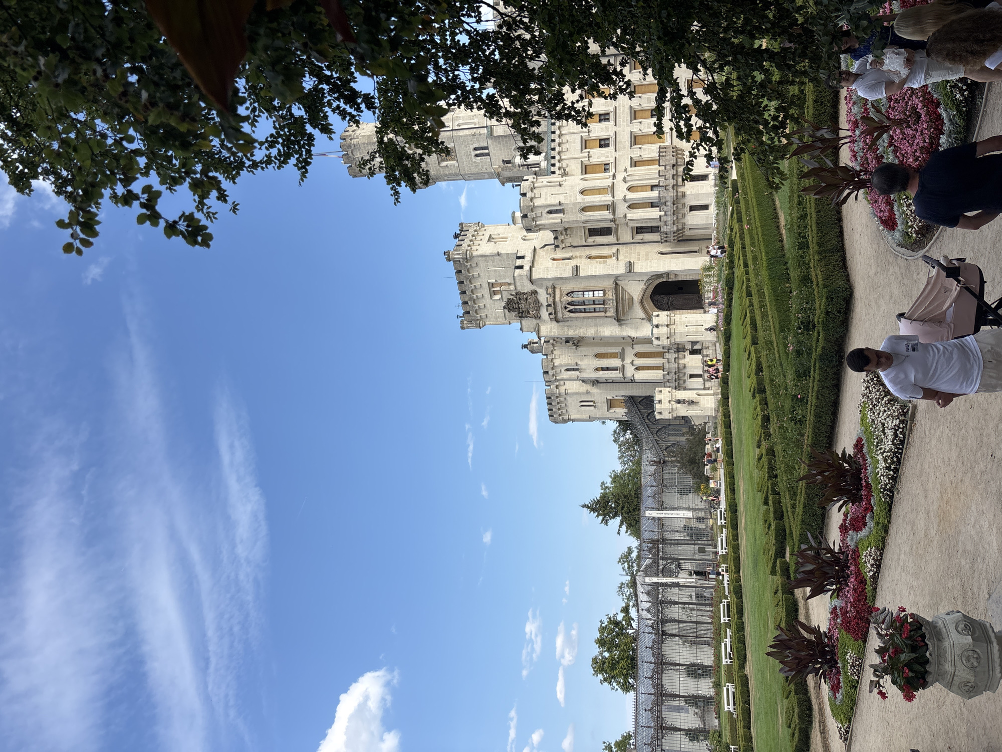 This is a photograph taken in Hluboká nad Vltavou, Czechia, featuring Hluboká Castle. The image shows a clear blue sky with wispy clouds. In the foreground, a manicured garden with flower beds and trimmed hedges is visible. A large urn with red flowers sits to the left. Several people are present, including a man in a white t-shirt pushing a stroller, another man in a black t-shirt, and a group of people further back in the garden, some of whom appear to be looking at the castle. In the middle ground, a long glass conservatory with white benches in front of it is situated. The prominent structure in the background is Hluboká Castle, a white building with multiple towers, turrets, and arched windows, displaying a neo-Gothic architectural style. The castle is partially obscured by the foliage of a large tree on the right side of the frame.