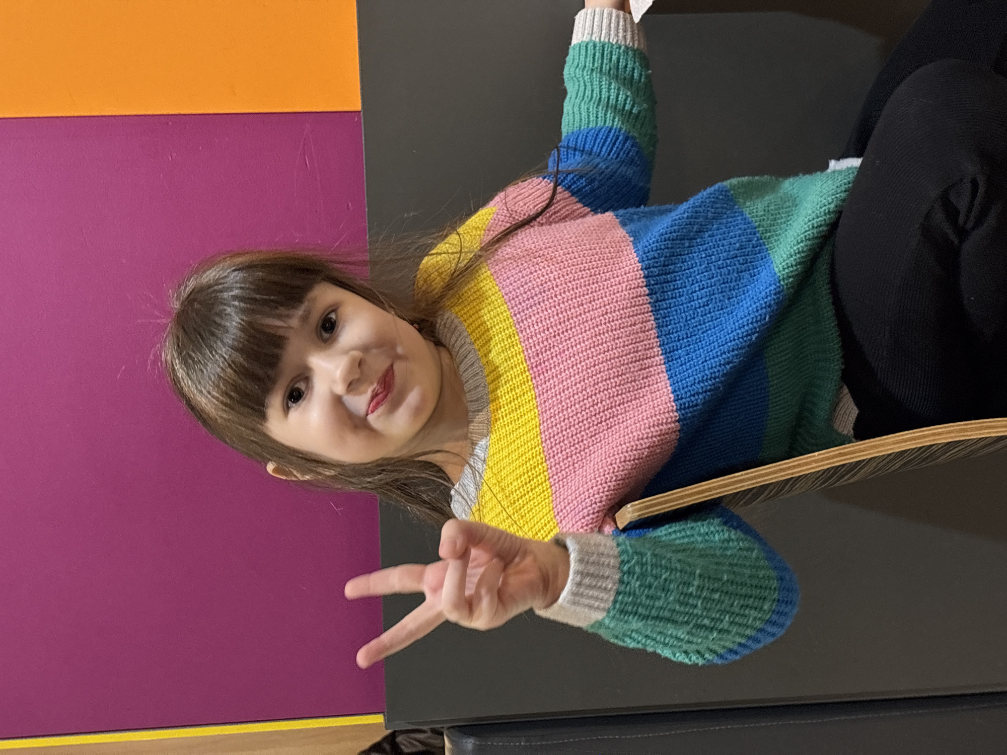 A young female child with dark hair and bangs is seated, looking directly forward with a smile. She is wearing a horizontally striped knitted sweater with sections of grey, yellow, pink, blue, and green, over black leggings. A red smudge is visible on her upper lip. Her left hand is raised, with the index and middle fingers extended to form a "V" sign. She is positioned in front of a brightly colored background consisting of a magenta wall panel, bordered by a yellow vertical stripe to the left and an orange vertical panel to the right. Below the wall panels, a dark grey padded bench is visible. Part of a wooden chair back is present to the right of the child. The specific location within the city of Izhevsk, Russia, cannot be determined from the visual evidence.