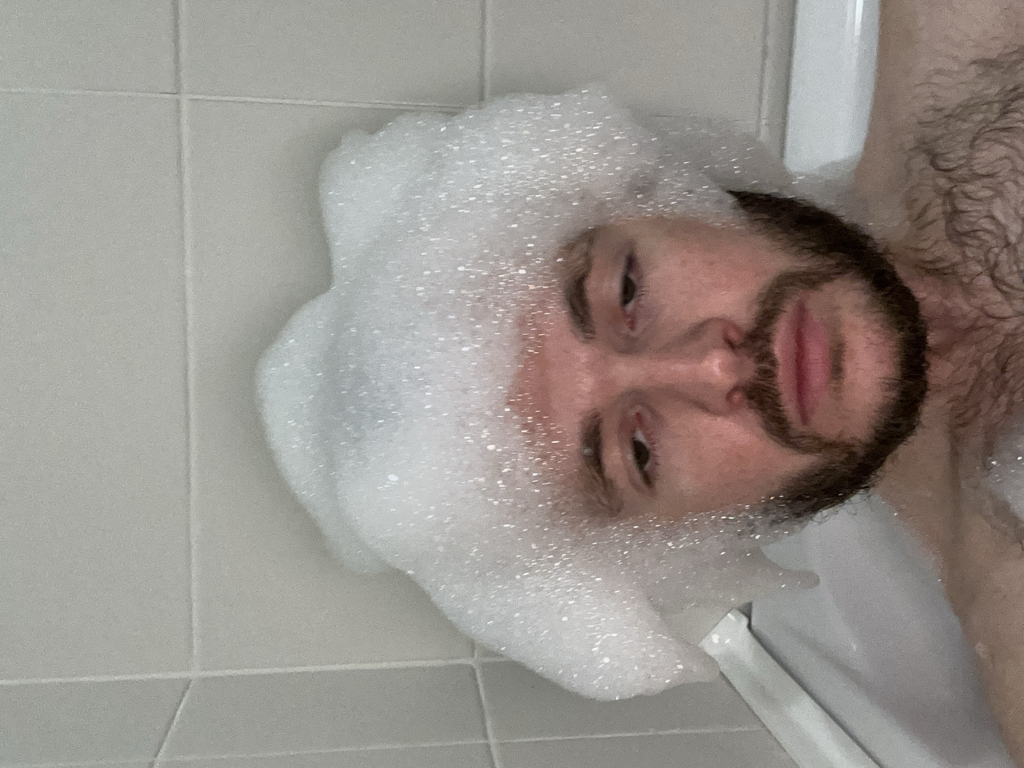 An overhead, sideways-oriented close-up photograph captures a person taking a bubble bath. The subject is a male with a beard and appears to have dark hair on his chest, which is visible on the right side of the frame. He is lying in a white bathtub, partially submerged in water, with a significant amount of white, frothy bubbles covering his head and neck area. His face, looking upwards towards the camera, shows a neutral expression, with his eyes open and lips slightly parted. The person's left eye is largely obscured by the foam, while his right eye is more visible. The background consists of light gray tiled walls, with grout lines clearly visible, indicating an indoor bathroom setting. A white rim of the bathtub is also visible at the bottom and right edge of the frame. The image is brightly lit, likely by artificial indoor light. No text is visible in the image.