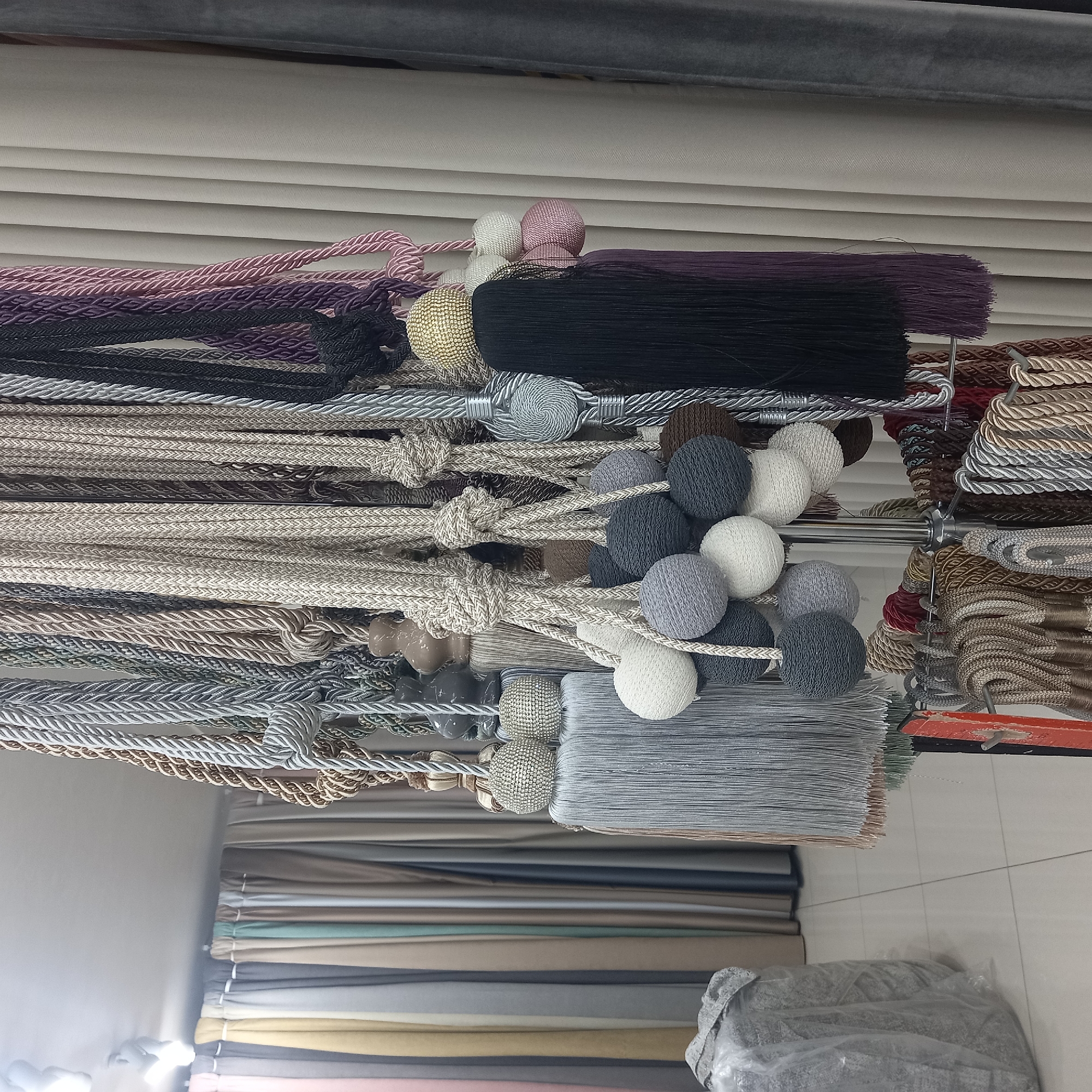 The image depicts a retail display of curtain tiebacks and tassels in a store. The tiebacks are made of braided cords in various colors including beige, grey, silver, black, and purple. Many of them are adorned with decorative elements such as knotted cords, tassels made of thin strands, and spherical ornaments covered in beads or woven with yarn. In the background, several draped curtains in muted tones of pink, yellow, grey, beige, and green are visible. The floor is tiled with large square tiles. The overall setting suggests a fabric or home decor store. No people are present in the image. The image was taken in Briceni, Moldova.