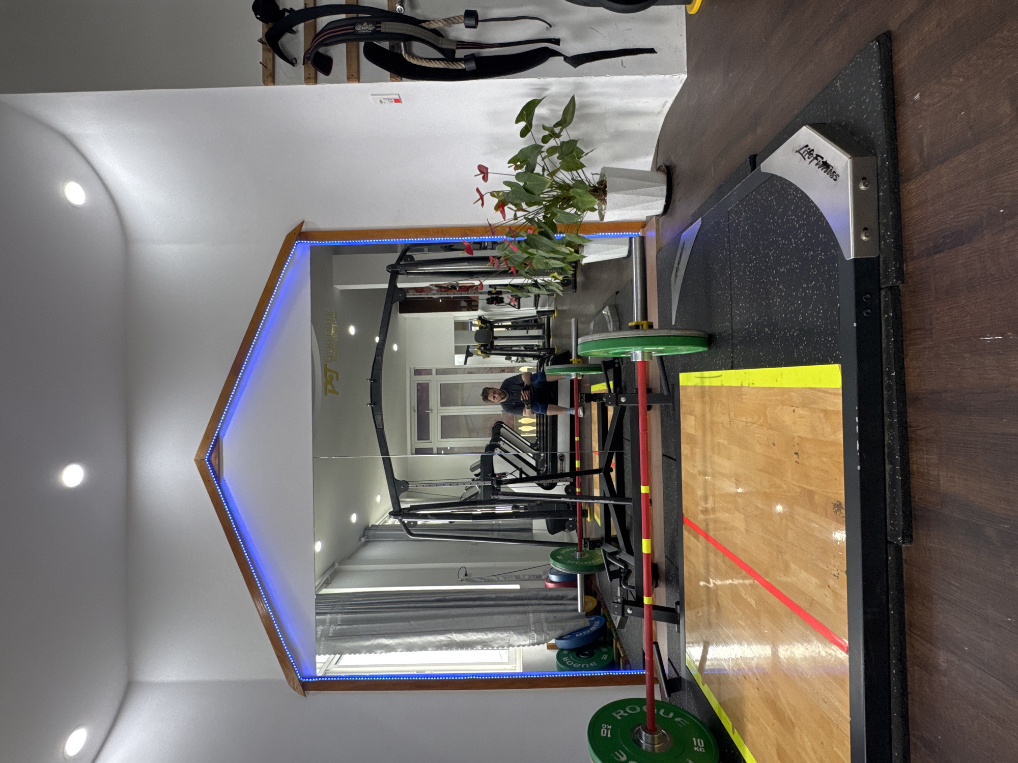 This image captures an indoor home gym or private fitness studio, with a provided context indicating its location in Da Lat, Vietnam. The room features white walls and a warm wooden floor.
A prominent feature is a large, custom-shaped mirror on the left, framed by wood and illuminated by a strip of blue LED lights. Reflected in the mirror is a man, possibly resting or observing, seated amidst gym equipment. The reflection also shows windows with curtains, suggesting natural light entering the room.
In the foreground and extending to the right, a dedicated weightlifting platform is visible. It comprises a central wooden section, marked with a distinctive red line and yellow border, flanked by textured black rubber matting. A barbell, loaded with green "ROGUE 10 KG" weight plates, is positioned on the platform. Additional blue weight plates, marked "SBD 10 KG," are stacked nearby. A metal bracket on the platform bears the "Life Fitness" logo. Part of a squat rack or power cage is also discernible.
Above the right side of the mirror, a wall-mounted rack displays several black weightlifting belts. A potted plant with striking red Anthurium flowers adds a splash of color to the space. The room is brightly lit by recessed ceiling lights, complemented by the ambient blue glow from the mirror's LEDs. The overall impression is of a clean, well-equipped, and inviting personal workout area.