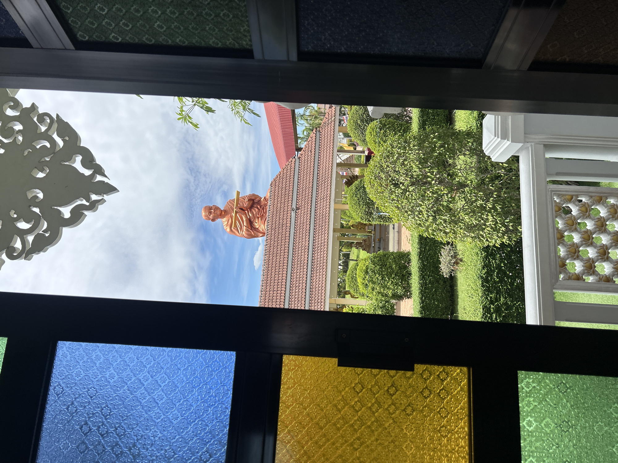 The image depicts a view through a stained-glass window onto a temple courtyard in Thai Ko Subdistrict, Thailand. In the foreground, colored glass panes in blue, yellow, and green are visible, framed by dark window trim. Beyond the window, a large copper-colored statue of a seated monk is perched atop a tiled roof structure. The courtyard features manicured hedges, a covered walkway with pillars, and more statues in the background. Palm trees and a red-roofed building are also present. The sky is partly cloudy. The scene is well-lit, suggesting it was taken during the daytime.