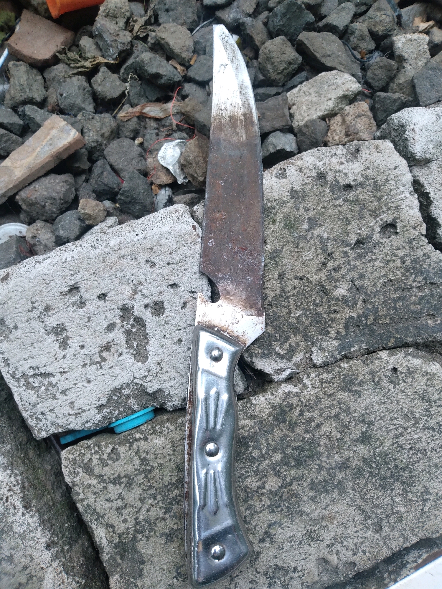 An overhead, medium shot shows a rusty, partially cleaned knife lying vertically on rough, gray concrete and gravel. The knife's blade is discolored with rust and has a white residue near the tip. The handle is made of polished metal with three rivets. The background consists of dark gray gravel, some broken concrete pieces, and a few bits of debris, including a small piece of red string. The scene appears to be an outdoor, possibly construction or debris-strewn area, located in South Tangerang, Indonesia.