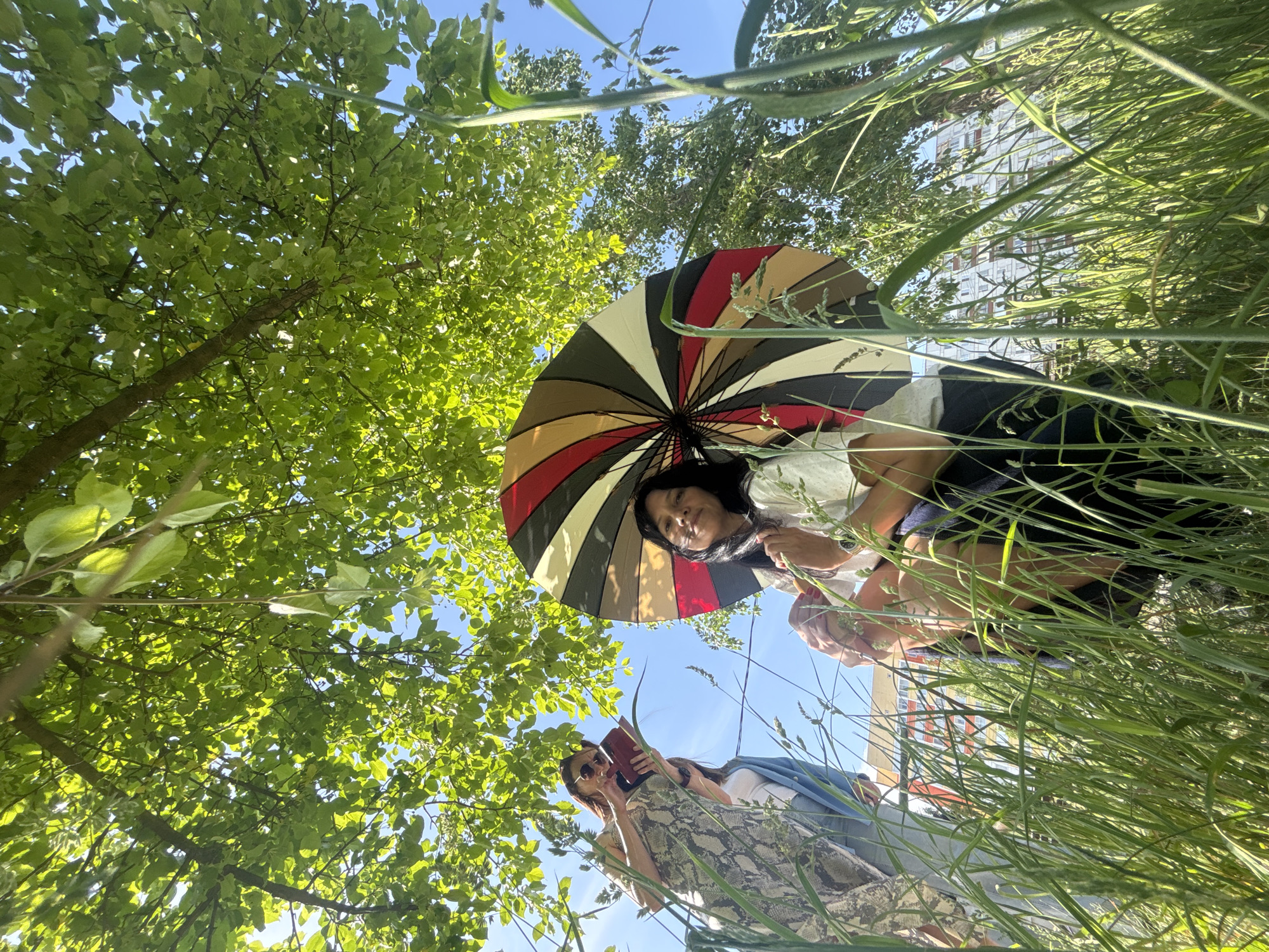 From a low angle amidst tall grass, two women are visible. One woman holds an open, multi-colored umbrella. The second woman is reading a red-covered book. Green trees are overhead against a blue sky. A building is visible in the background. This scene is in the city of Chaikovsky, Russia.