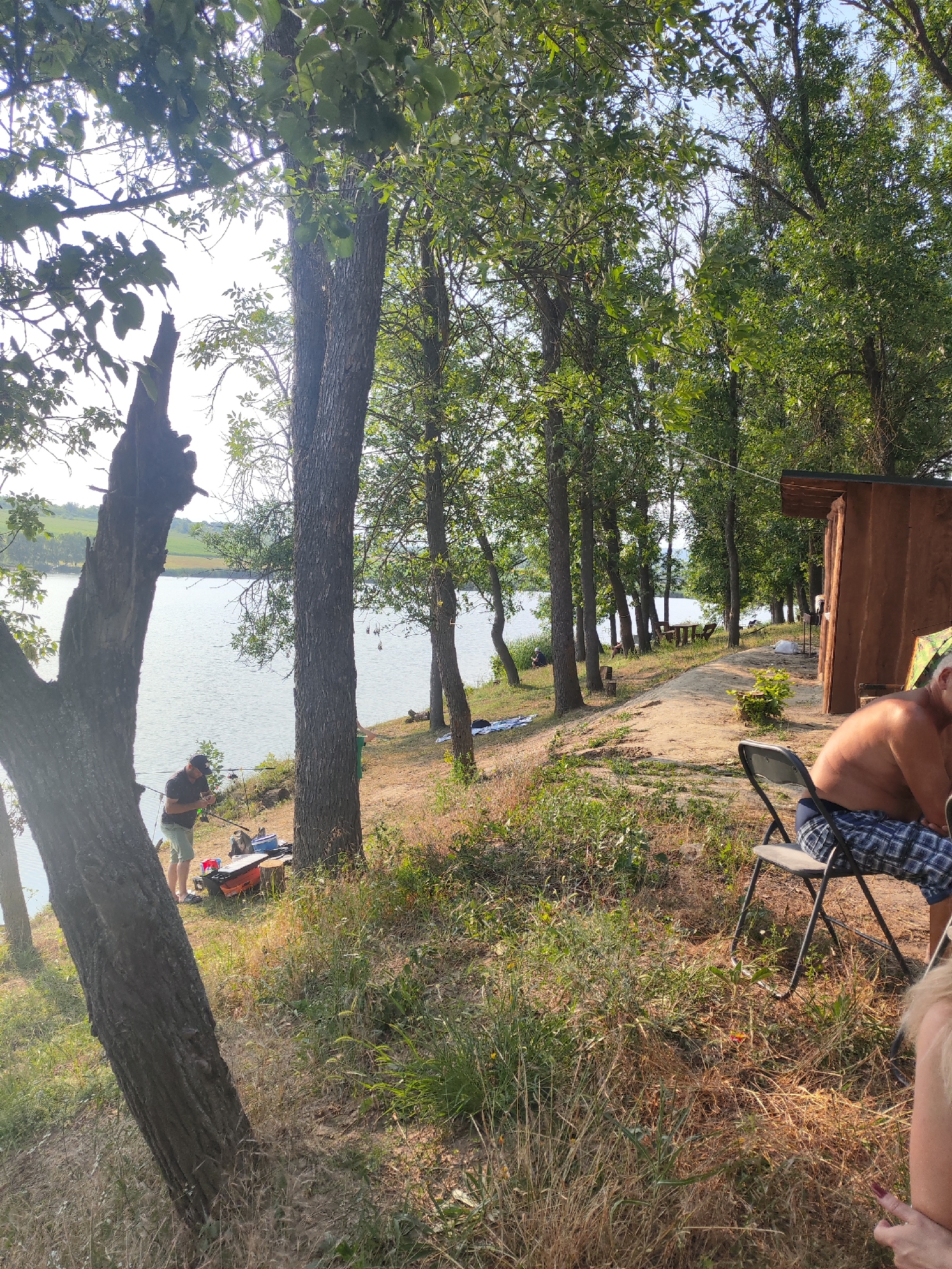 A daytime, outdoor scene depicts a body of water, likely a lake, on the left side of the image, with a grassy bank and several trees lining the shore. A person in a black cap and shorts is standing near the water's edge, appearing to be fishing, with fishing rods and tackle boxes visible on the ground. Further back, a small wooden structure, possibly a shed or cabin, is situated on the right side of the frame. A man wearing plaid shorts is seated on a folding chair in the foreground, looking away from the camera. The environment suggests a natural setting, possibly a recreational area near Găleștii Noi, Moldova, with lush green foliage dominating the background.