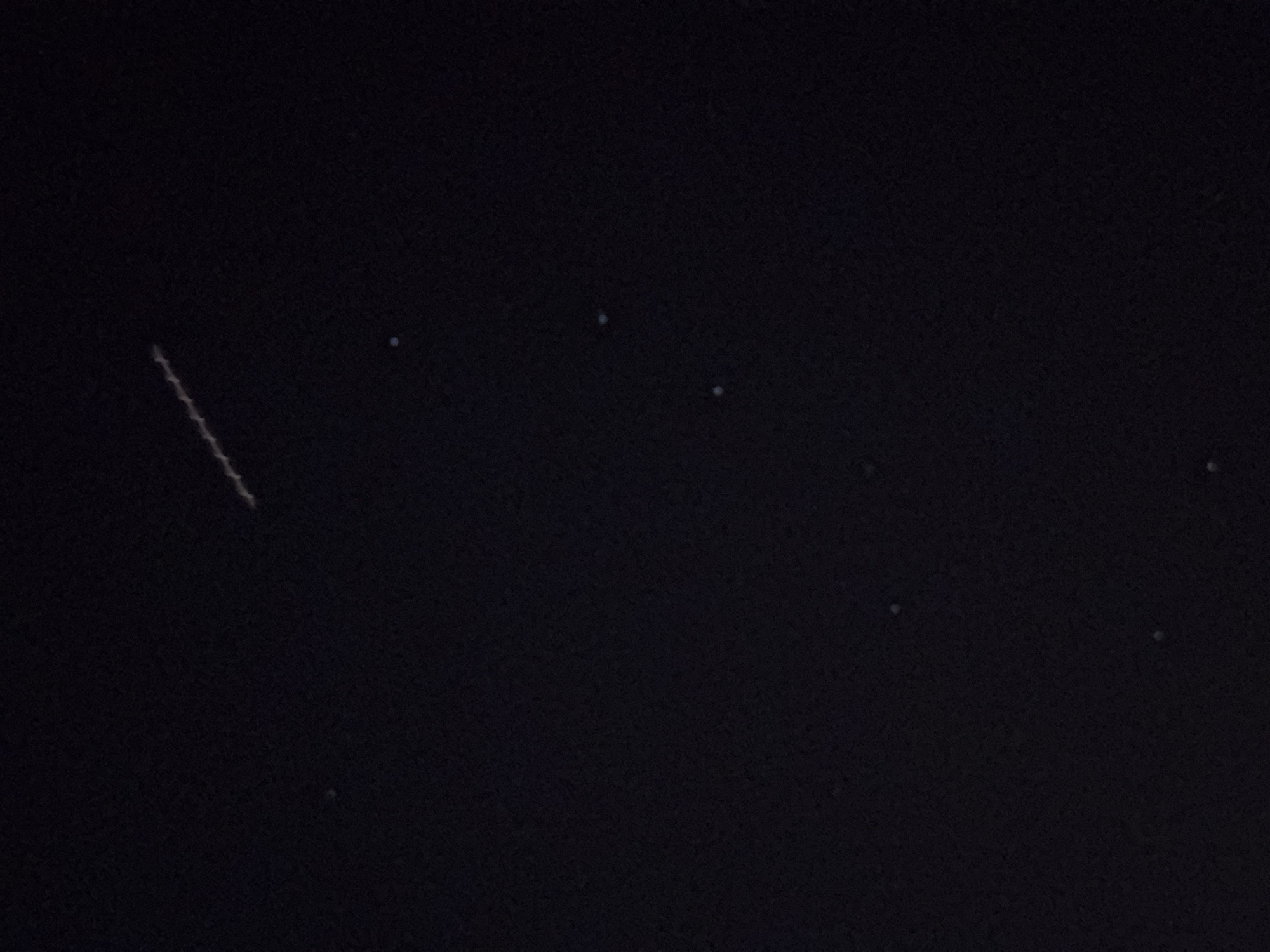 The image is a nighttime photograph of the sky above Shkorpilovtsi, Bulgaria. The sky is dark, with a few scattered stars visible. A trail of lights, likely from a satellite or aircraft, is visible diagonally across the upper portion of the image. The trail is composed of small, distinct lights. No other objects, people, or structures are discernible in the frame. The environment appears to be an outdoor, unlit area, consistent with a clear night sky.