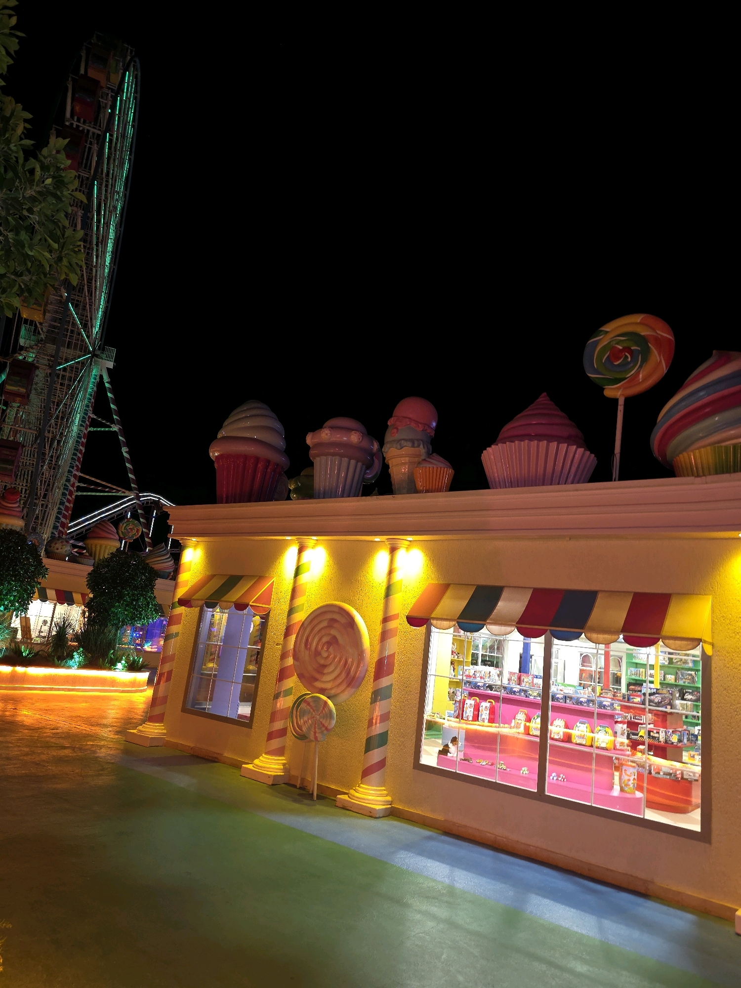 The image depicts a whimsical, candy-themed facade of a building, likely a toy or candy store, at night. The building is painted a bright yellow and adorned with oversized, colorful candy and cupcake decorations on its rooftop. Two large lollipops, one on a stick and another embedded in the facade, add to the confectionery theme. The storefront features large windows displaying shelves filled with toys or candy. Striped awnings in red, yellow, and blue cover the windows, and decorative columns with red and white stripes flank the entrance.
To the left of the building, a large Ferris wheel, illuminated with green lights, stands prominently against the dark night sky. The Ferris wheel is a significant landmark, suggesting this location is an entertainment or amusement area. The ground in front of the building appears to be a paved walkway or plaza, with areas of green and blue surfacing. The overall ambiance is festive and playful, designed to attract visitors, especially families. This scene is located in Bodrum, Turkey, a popular tourist destination known for its vibrant nightlife and entertainment options.