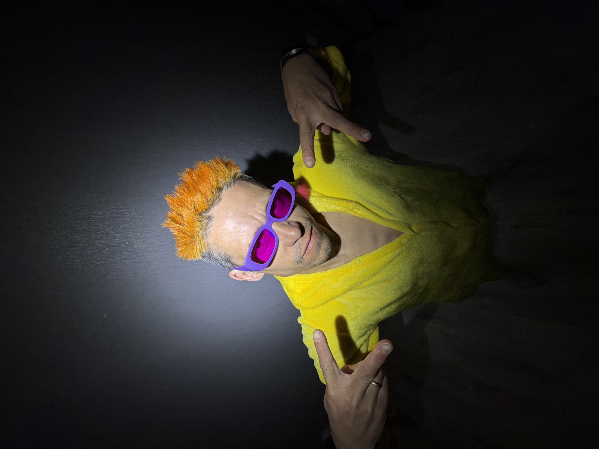 An adult male is prominently featured in a dramatic, top-down shot, positioned diagonally against a dark, textured background. He sports striking spiked hair, dyed a vibrant orange-yellow with visible gray roots. His eyes are obscured by bright purple, rectangular sunglasses, and he maintains a neutral to slightly smirking expression while looking towards the camera.
He is dressed in a yellow, textured garment, possibly a robe or a hooded top. His hands are visible, one above his head extending two fingers (index and middle), and the other closer to his chest forming a "rock on" or "devil horns" gesture. A silver-toned watch is worn on his right wrist, and a simple band ring is on the ring finger of his left hand.
The scene is lit by a single, strong light source from the top-left, creating sharp highlights on the subject's hair, face, and clothing, and casting deep, defined shadows across the dark background. This directional lighting enhances the textures and creates a high-contrast, stylized portrait. No discernible text is present in the image.