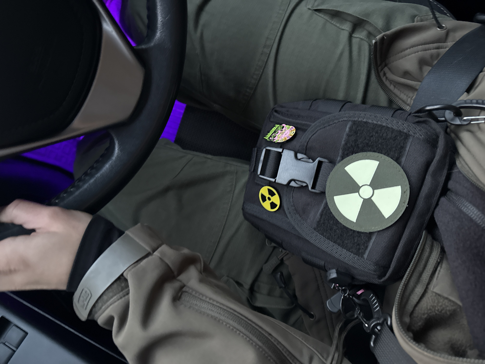 A person sits in the driver's seat of a vehicle, possibly a car, with a black tactical bag resting on their lap. The bag is adorned with a radiation symbol patch and a yellow radiation hazard pin. The person is wearing olive green clothing, including tactical pants and a jacket with a visible zipper. A black steering wheel is visible on the left, with a purple light emitting from the dashboard area. The scene is likely indoors, inside the vehicle, but there is no indication of the location being Kyiv, Ukraine, or any other specific city.