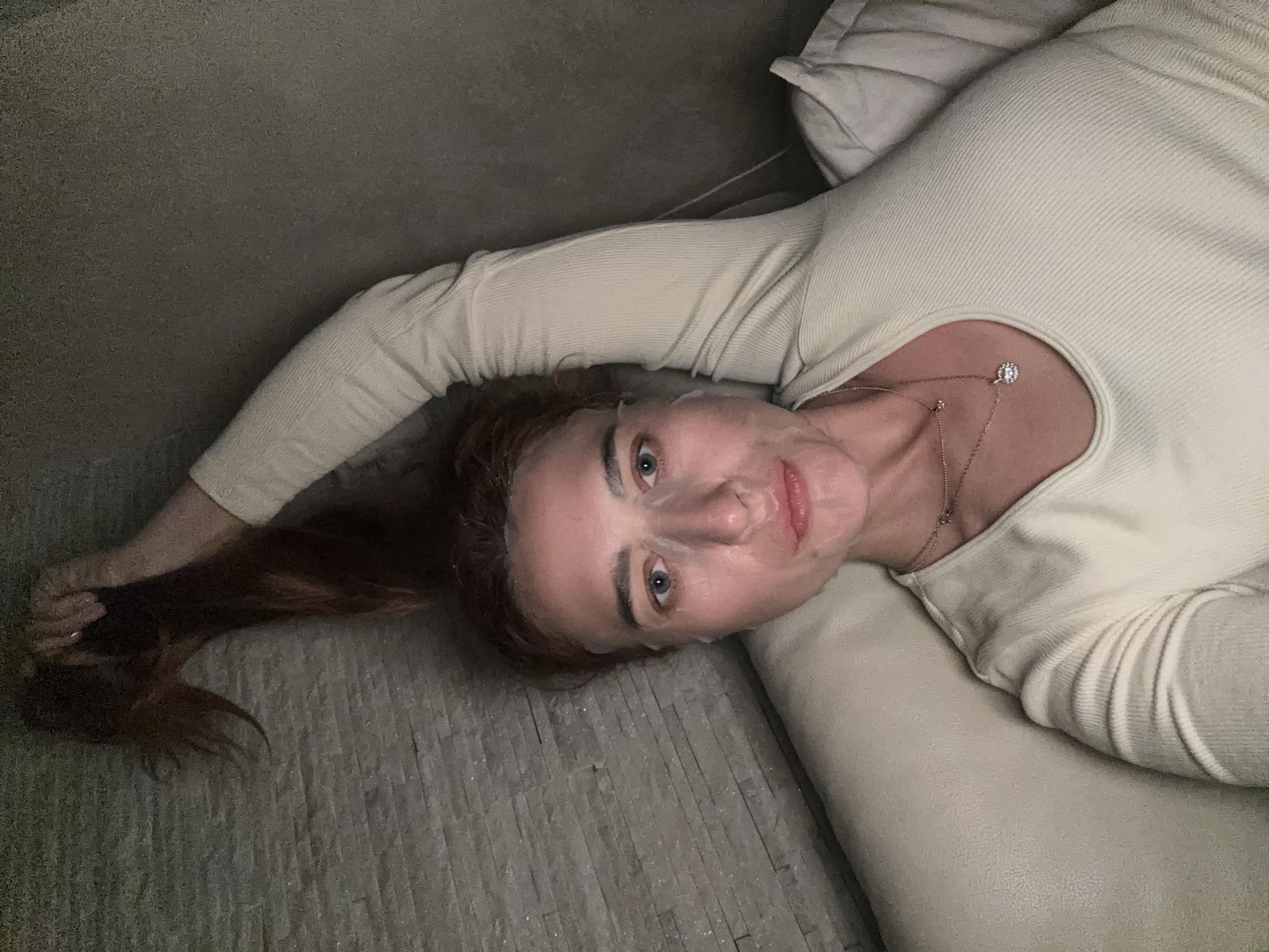 A woman lies on a light-colored upholstered surface, possibly a couch or daybed, her head tilted to the right. She wears a light beige, long-sleeved ribbed shirt, a necklace with a pendant, and what appears to be a sheet mask on her face. Her reddish-brown hair is pulled to the left, and she is holding the end of it in her hand. The background features a gray textured wall and a pillow behind her head. The location cannot be determined from the provided image. The image is focused on the woman's face and upper body.