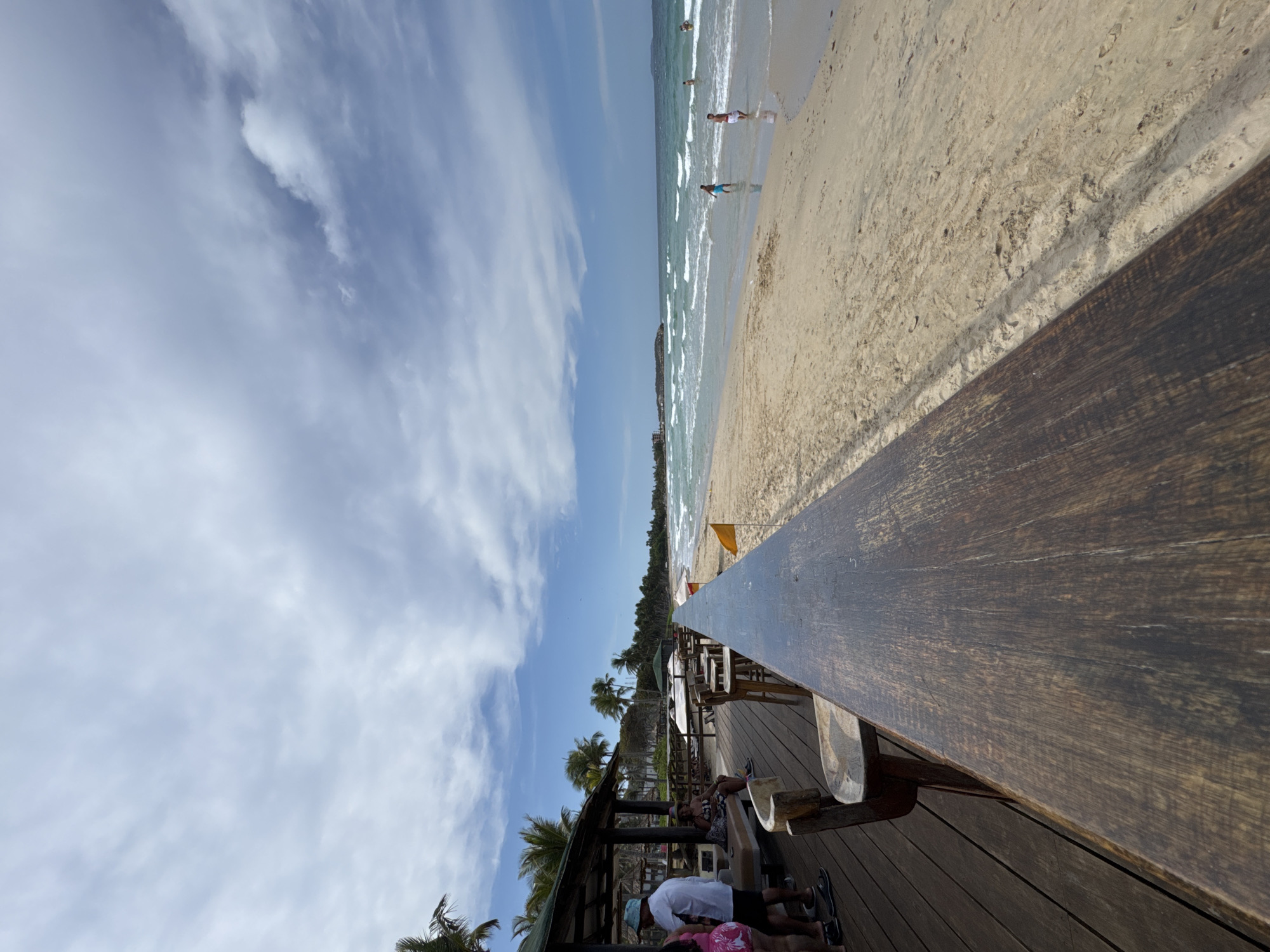 The image captures a vibrant, sunny day at a beachside location in El Tirano, Venezuela. The scene is divided horizontally, with a wide expanse of blue sky and white, wispy clouds at the top, a blue-green ocean with gentle waves in the middle, and a light-colored sandy beach to the right.
In the foreground and midground on the left, there's a wooden deck area, likely part of a restaurant or bar, positioned elevated above the beach. A long, dark wooden railing runs diagonally across the image from the bottom right towards the center, separating the deck from the sandy beach. On the deck, several people are present, some sitting at tables under a shaded structure, others standing. Palm trees are visible throughout the deck area, contributing to a tropical ambiance.
Several individuals are enjoying the water on the beach, seen wading or swimming near the shore. One orange flag is planted in the sand on the beach. The overall activity suggests leisure and relaxation, with people enjoying the beach and the amenities of the establishment. The clear blue sky and bright sunlight indicate daytime, likely mid-day, with pleasant weather conditions. No discernible text is visible in the image.