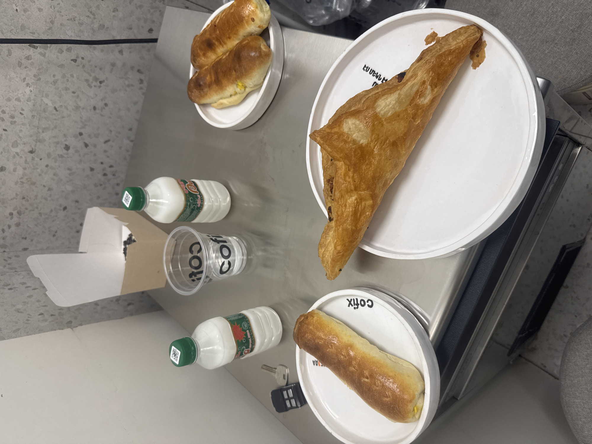 The image depicts a selection of food and beverages on a stainless steel surface. There are three white plates, two containing sausage rolls, and one with a pastry. Additionally, there are two small white bottles, a plastic cup, and a brown paper bag with contents. The presence of the "co fix" branding on the cup and one of the plates suggests this is a setting related to the Coffeebox brand, which is a coffee shop chain. The items are arranged on what appears to be a counter or table. A set of keys and a car fob are also visible. While it is not possible to pinpoint the exact location within Yerevan, Armenia, based solely on this image, the presence of Coffeebox branding strongly suggests the scene is within a Coffeebox outlet in the city. No people are visible in the image.