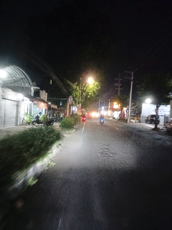 The image captures a night-time street scene in Mojosari, Indonesia, likely taken from a moving vehicle or one with its headlights on, illuminating the immediate foreground. The asphalt road, which appears somewhat textured or reflective, stretches into the distance, with visible ripples or patterns in the illuminated section.
Several motorcycles are present: at least two are parked on the left sidewalk near buildings, while two more, one with a red tail light and another with a blue headlight, are moving away from the camera further down the road. Buildings line both sides of the street. On the left, several storefronts are visible, some with closed roller shutters and others open, featuring arched corrugated metal canopies and potted plants. The right side also shows commercial establishments indicated by illuminated signs.
Streetlights illuminate the scene, with a prominent yellowish-orange streetlight on the left casting light on nearby tree leaves. Utility poles with numerous overhead wires are visible on the right. Bushes and trees line the sidewalks, with the foreground vegetation on the left appearing blurred due to motion.
A circular traffic sign with a white circle, red border, and a horizontal white bar (commonly indicating "No Entry" or a similar prohibition) is visible on the left sidewalk, partially obscured by foliage. Another white sign on the right side of the road has illegible text. The overall atmosphere is that of a bustling local street after dark.