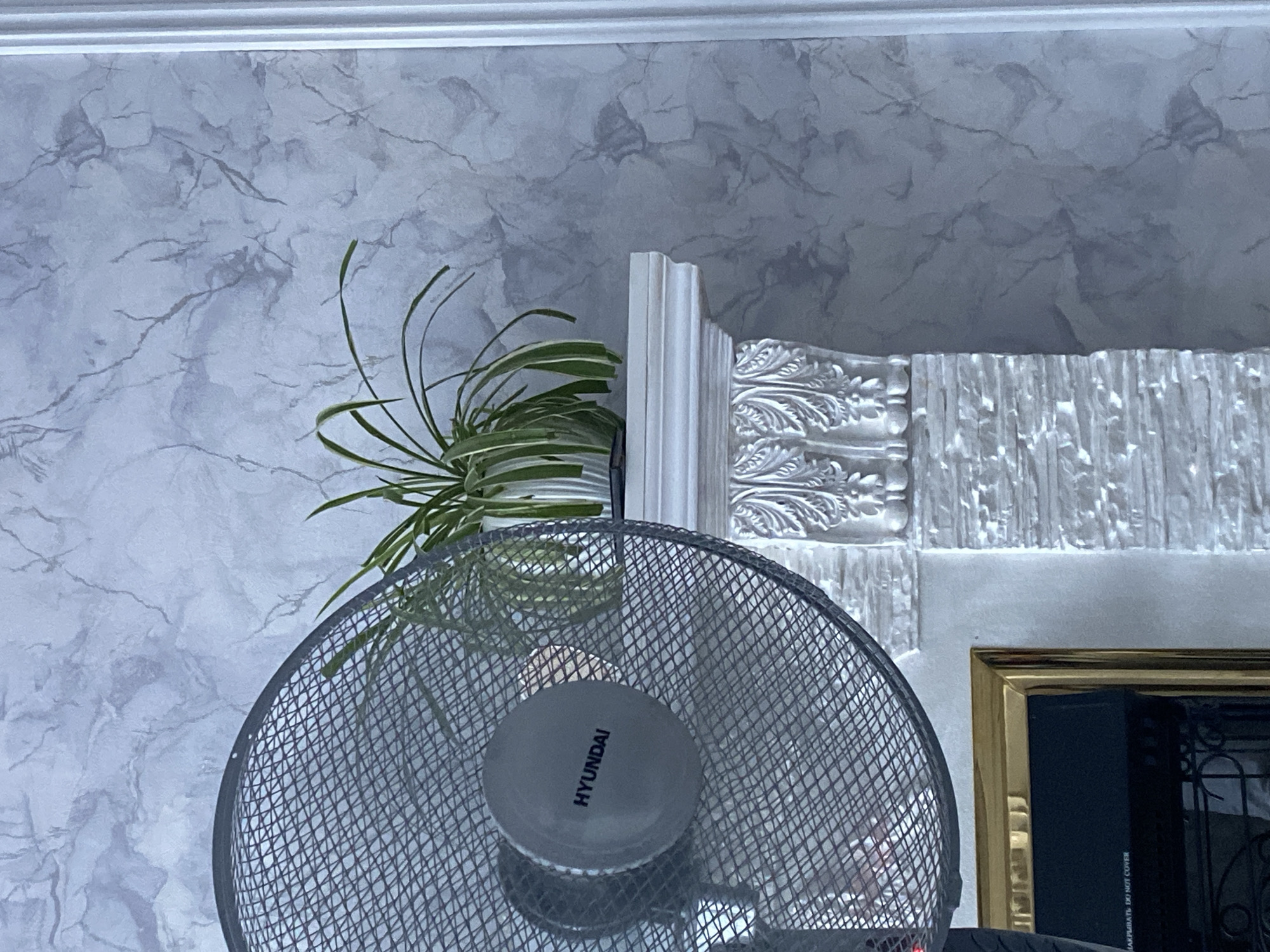 The image shows a close-up of a fan with the brand name "HYUNDAI" visible on its central hub, positioned in front of a fireplace mantel. The mantel is white and features decorative carvings. On top of the mantel sits a potted plant with long, green leaves. The background is a wall with a marbled texture in shades of grey and white. The bottom left of the image shows part of what appears to be an electric fireplace with a golden frame and a black insert containing a warning label that reads "НАКРЫВАТЬ DO NOT COVER". The scene appears to be an indoor setting, likely a living room, possibly in Uchaly, Russia, based on the provided context. No people are visible in the image.