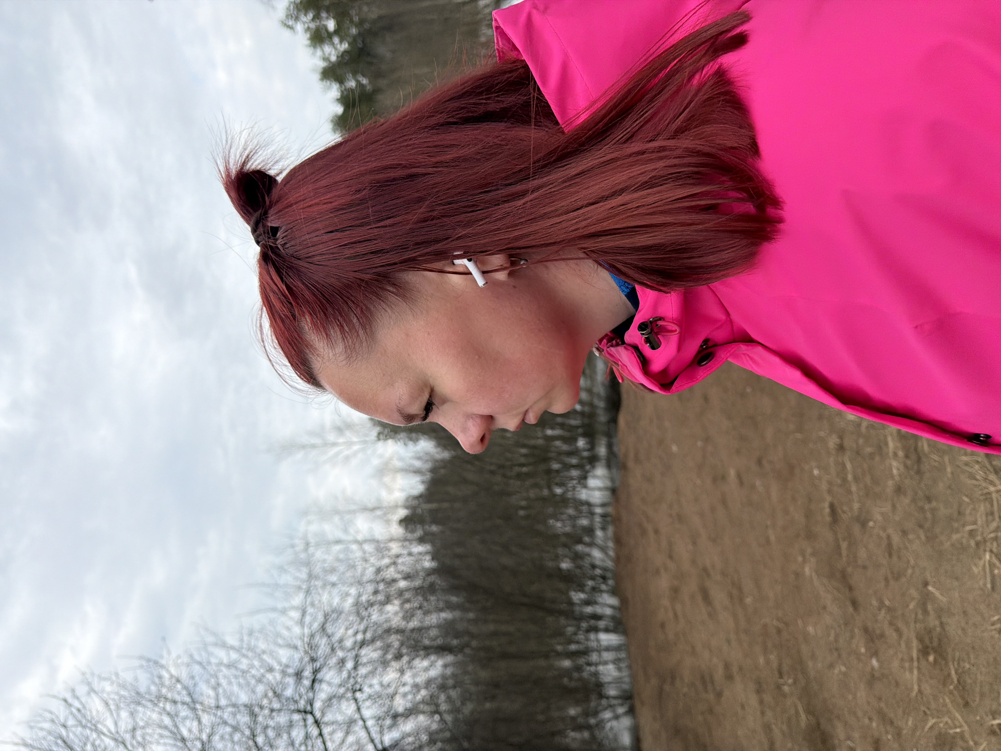 A woman stands outdoors on a cloudy day. Her dark red hair is pulled back into a small bun, with loose strands framing her face. She is wearing an earbud in her left ear and a bright pink jacket. The background is a mix of brown grassy ground and leafless trees stretching toward a cloudy sky. This photo may be taken in Chaikovsky, Russia.