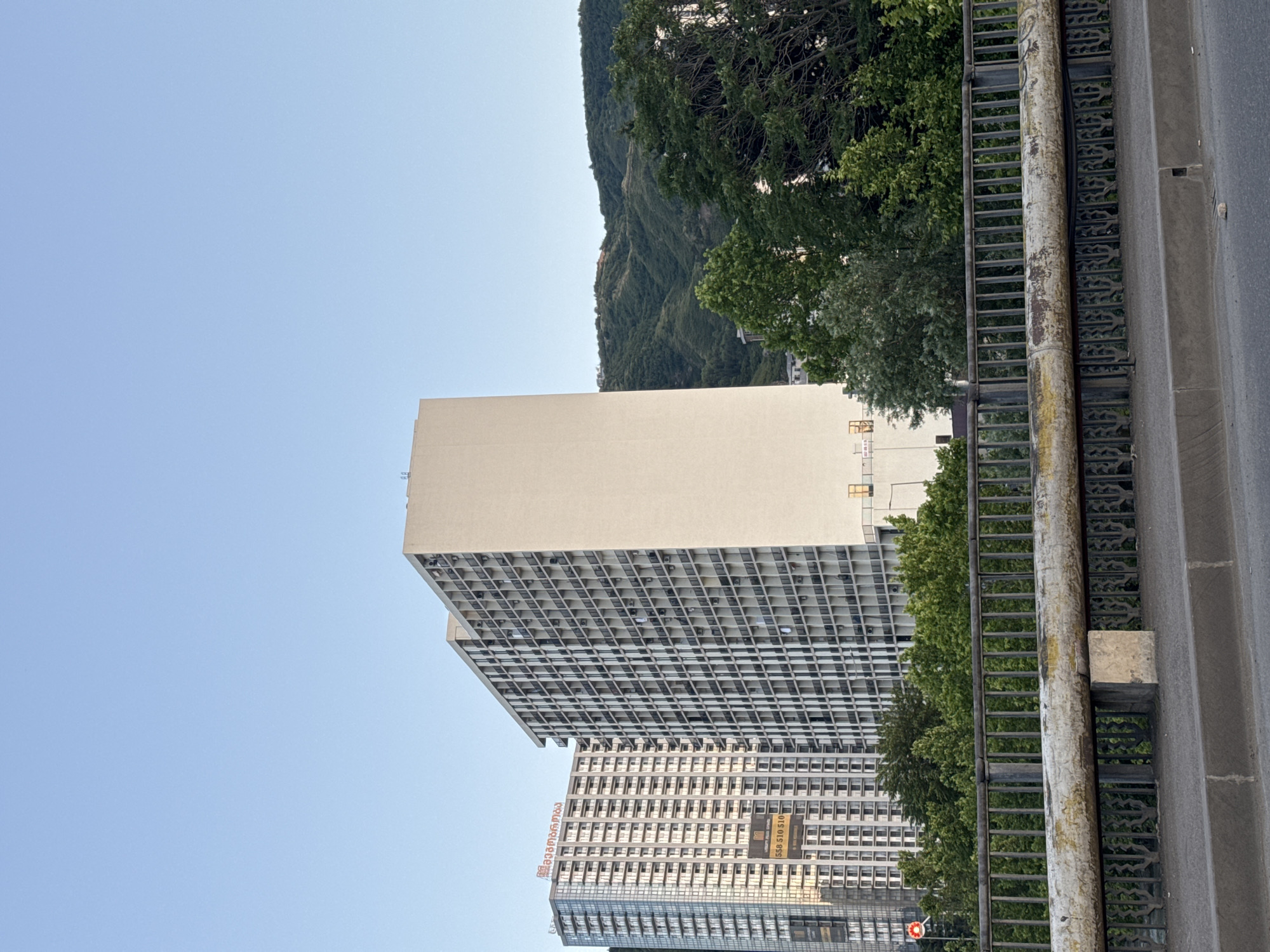 A full shot shows two residential apartment buildings in Tbilisi, Georgia. The building on the left is taller and has a glass facade, while the building on the right is shorter and has a white facade with many balconies. Both buildings are surrounded by trees and a green hillside. In the foreground, there is a bridge with a metal railing. The sky is clear and blue.