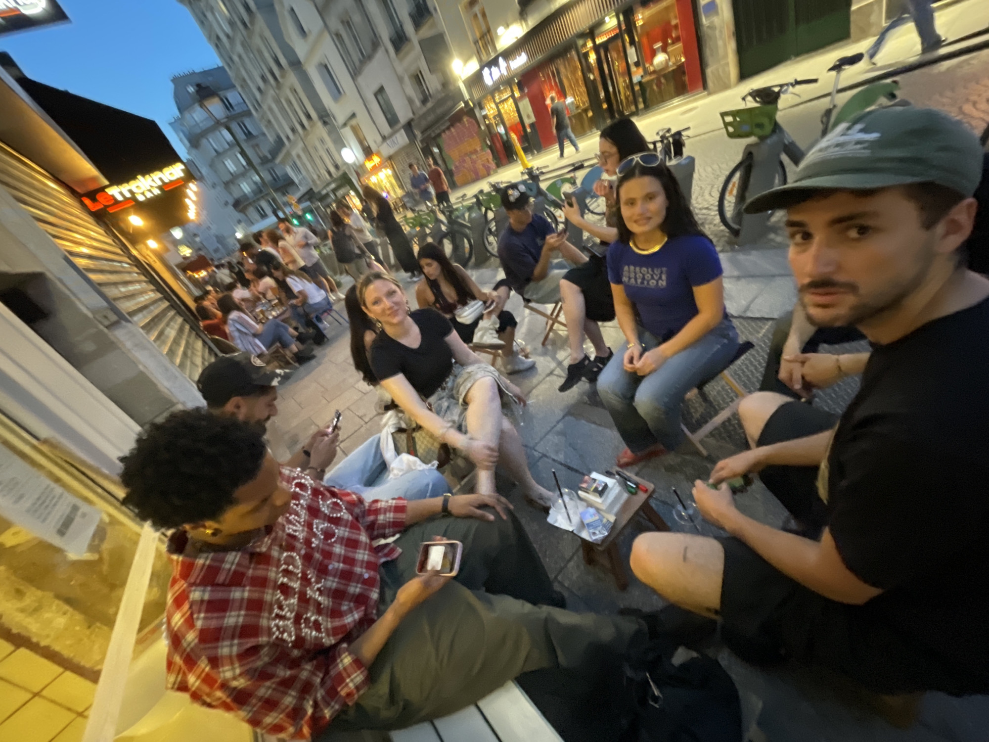 The image shows a group of people seated outside a bar in Paris, France. The bar, named "Le Trakhar Bar", has a red and orange sign. Several individuals are seated on outdoor chairs, engaged in conversation and interaction. One person in the foreground is wearing a green cap and a black t-shirt, looking towards the camera. Another person in the lower left wears a red plaid shirt with embellishments. Several others are scattered around the area, with some looking at their phones. Bicycles are parked nearby. The environment indicates an urban setting with buildings lining the street, typical of Paris. The lighting suggests it is dusk or evening.