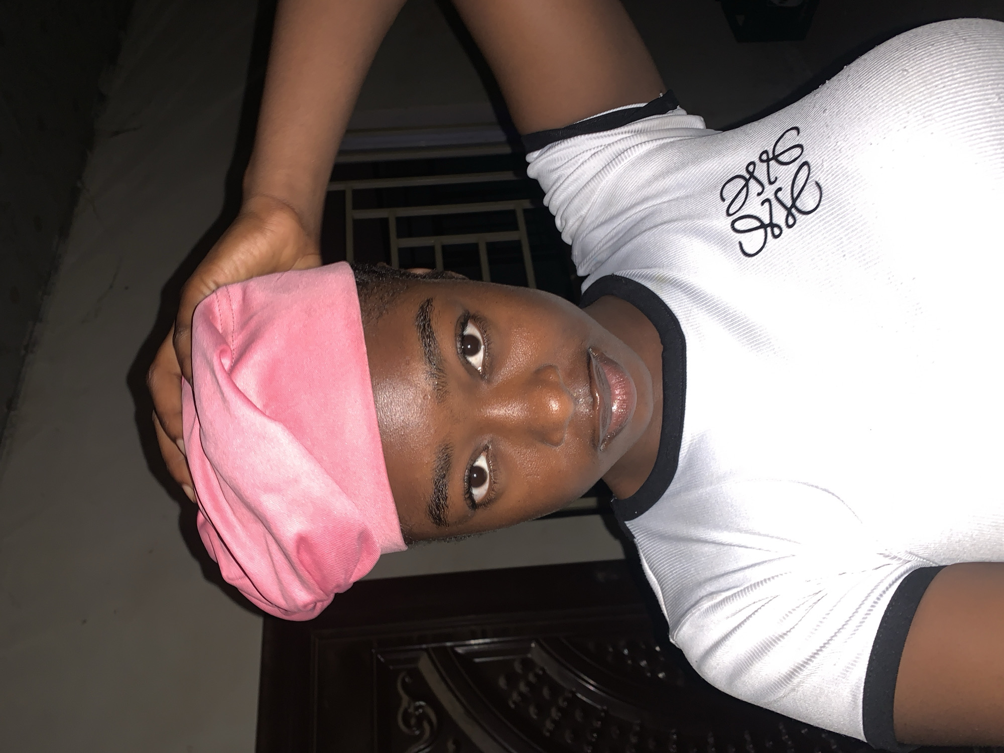 The photograph depicts a person with dark skin wearing a pink head covering and a white t-shirt with black trim. The t-shirt features a dark emblem on the upper chest. The individual is looking directly at the camera. The background is indistinct but appears to be indoors and dimly lit. The location is the city of New Stadium, Nigeria.