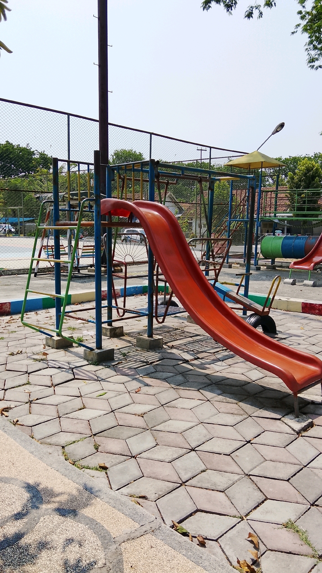 The image depicts a playground located in Tlogobendung, Indonesia. The playground features a prominent orange slide with a blue metal frame. To the left of the slide, there is a green and blue ladder. In the background, a chain-link fence encloses the area, with trees and buildings visible beyond. The ground is paved with hexagonal gray tiles, and fallen leaves are scattered across the surface. Several metal poles and other playground equipment, including what appears to be a seesaw made from a tire and metal, are present. A lamppost with a yellow umbrella-like shade is also visible. The sky is clear and pale.