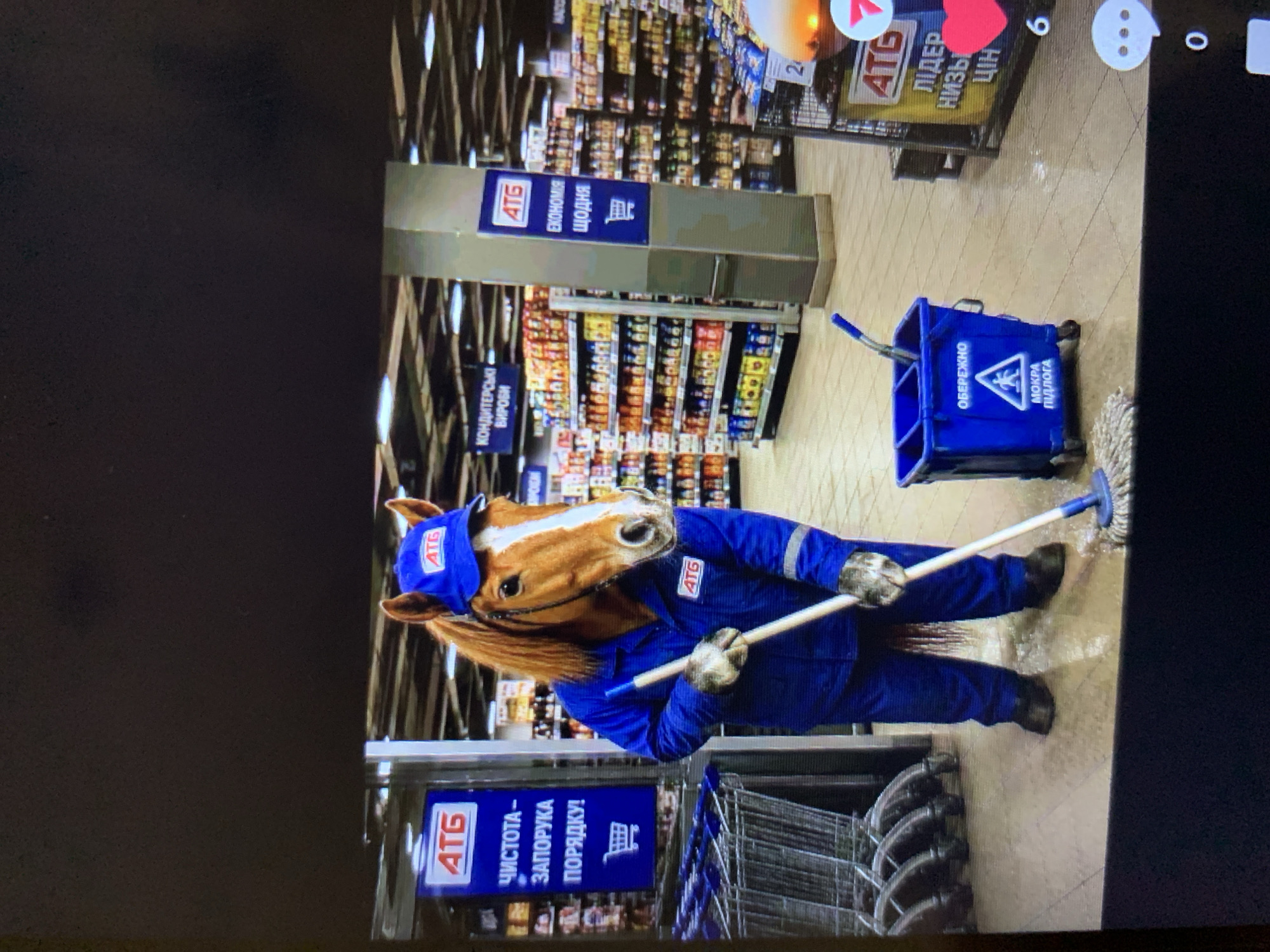 A man wearing a horse mask and a blue uniform with an "ATG" logo is mopping the floor of a supermarket. The supermarket appears to be an "ATB" store, indicated by the signage. The man is also wearing a blue baseball cap with the "ATG" logo. He is holding a mop and bucket. The floor is wet. There are shopping carts in the background. The image is taken from a slightly elevated angle.