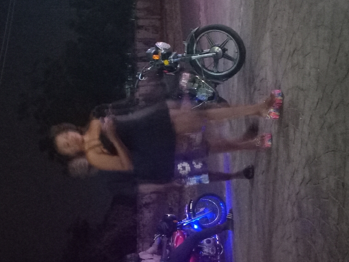 A blurry, dimly lit image shows a person standing near two parked motorcycles. The person, wearing a dark top and shorts, stands close to the motorcycles. One motorcycle is at the top of the image and the other is at the bottom, which has blue lights. The background suggests a night scene or a poorly illuminated area. The image appears out of focus, obscuring finer details.